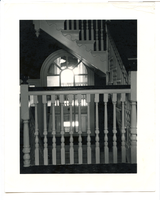 Black and white photograph of white banister, window in background shows staircase on other side of house.