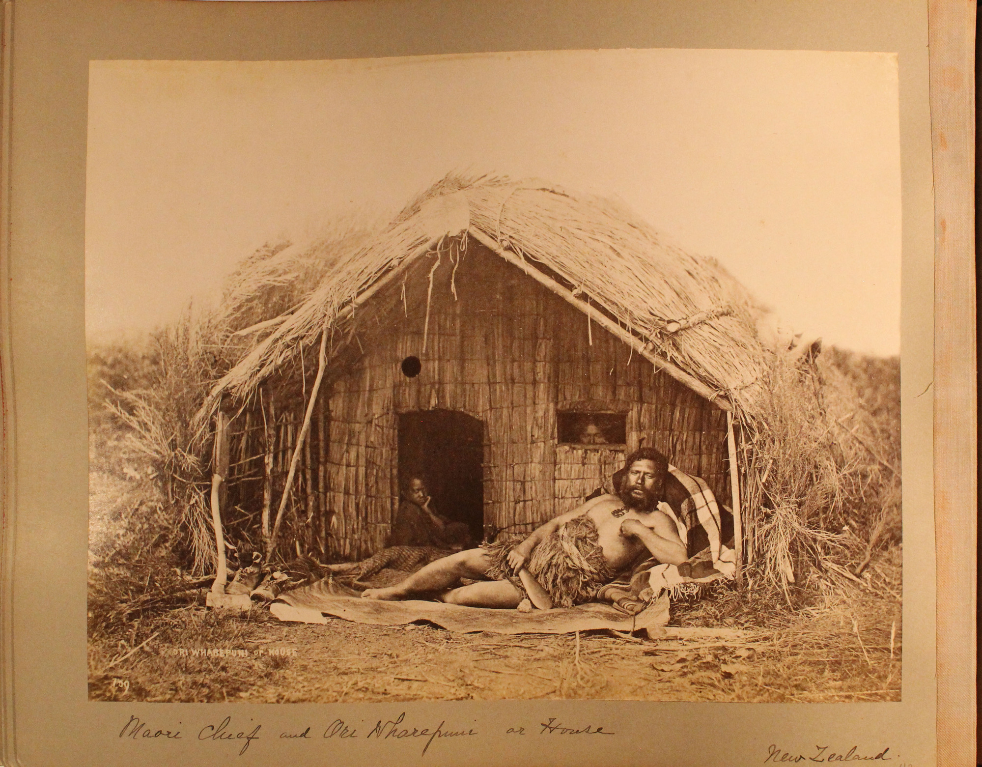 A Maori man reclines in front of a small reed house. He wears a fringed garment around his middle and holds a small wooden paddle. A boy sits in the doorway while an adult peers out of a small window.