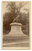 Photograph of a life-size or larger statue of Henry Wadsworth Longfellow. Statue is on a tall pedestal outdoors. Wooden house in the background. Longfellow wears a suit and is draped in some kind of cloth, possibly a professor/graduation gown. He has chin-length hair and a full beard.