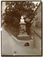 Henry Wadsworth Longfellow statue situated in triangular outdoor plaza. Plaza has grass and trees. Photograph shot from the statue's front. Building in the background on the photograph's right.