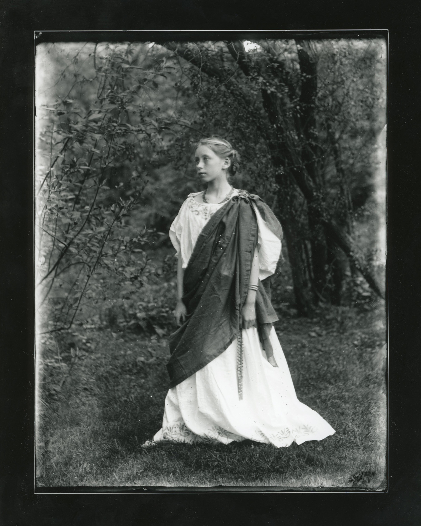 Young white woman stands with heavy shawl fastened at shoulder outdoors. Outfit looks Grecian.