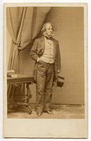 Full-length view of Henry Wadsworth Longfellow standing. He holds his hat in his left hand by his side and puts his right hand in his pants pocket. On the left side of the image are billowing drapery and a Renaissance Revival table with a book and indistinguishable object. Longfellow is wearing a light, striped vest, and has graying sideburns.
