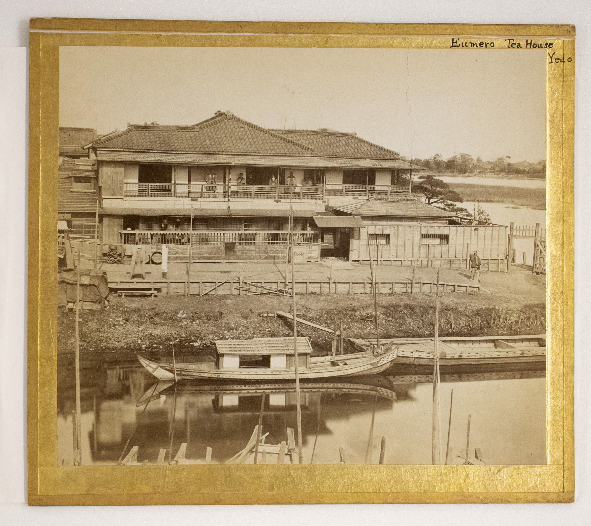 Sepia photograph representing Japanese buildings with people standing on the balcony and in front, boats in water are photographed in the foreground.