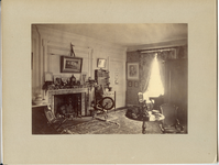 Black and white photograph of Victorian bedroom with fireplace, spinning wheel and bookshelf featured.