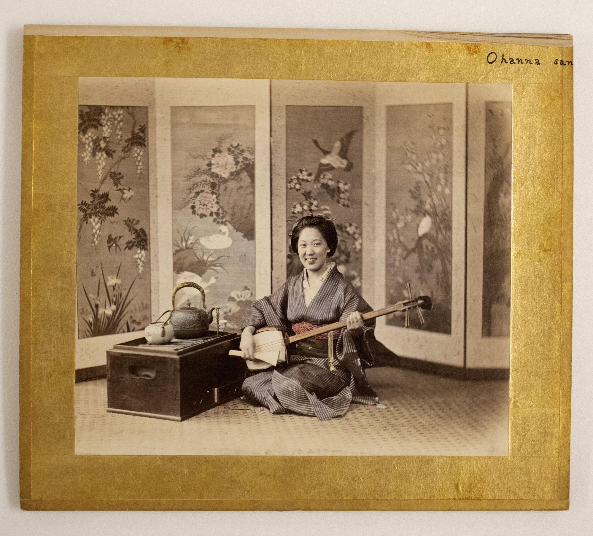 Sepia photograph with colored-in blue and red details representing a Japanese woman sitting on the floor next to a box with a tea set and holding a musical instrument. She is photographed with a folding screen behind her showing birds and plants.
