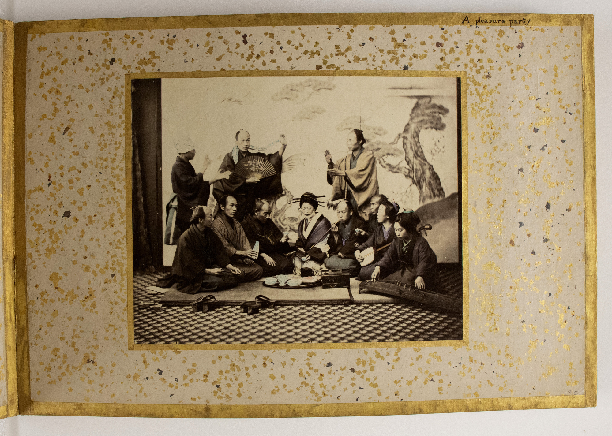 Sepia photograph colored with blue, yellow and purple shades representing a group of eleven Japanese men and women sitting on a mat and standing, holding musical instruments, fans, and pipes. They are photographed against a background with trees. The photograph is placed in the center of an album page decorated with a gold-leaf frame, colorful dots, and an inscription.