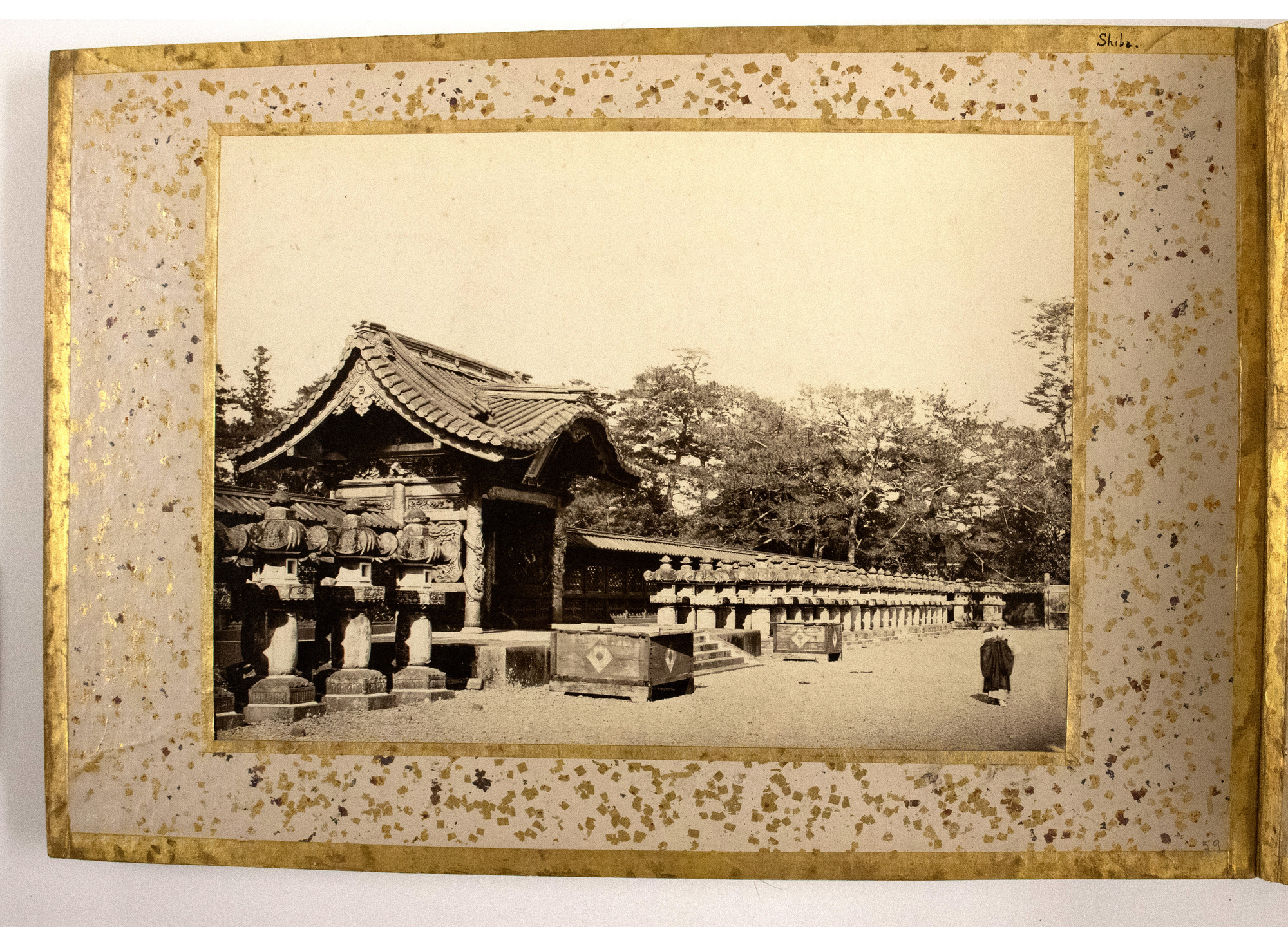 Sepia photograph representing a small Japanese building in the middle of a stone fence, possibly a gatehouse, with a row of stone lanterns along the fence and two boxes by the entrance, trees in the background and a person on the front right. The photograph is placed in the center of an album page decorated with a gold-leaf frame, colorful dots, and an inscription.