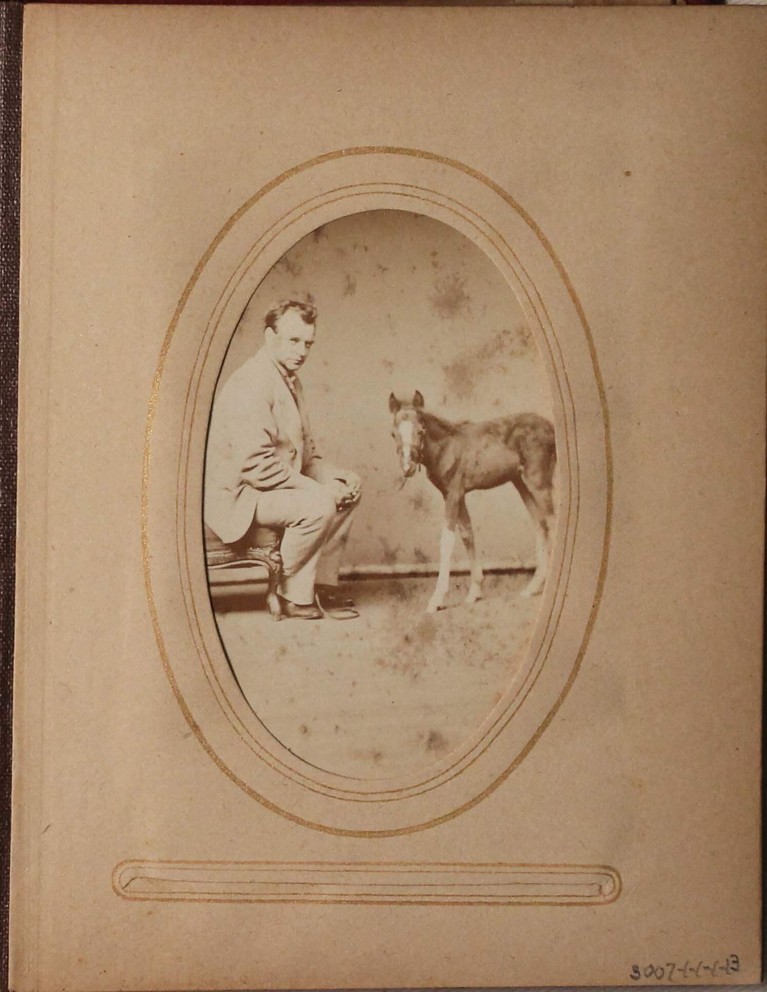 Black and white photograph of man seated looking at the viewer, across from him is a small horse.