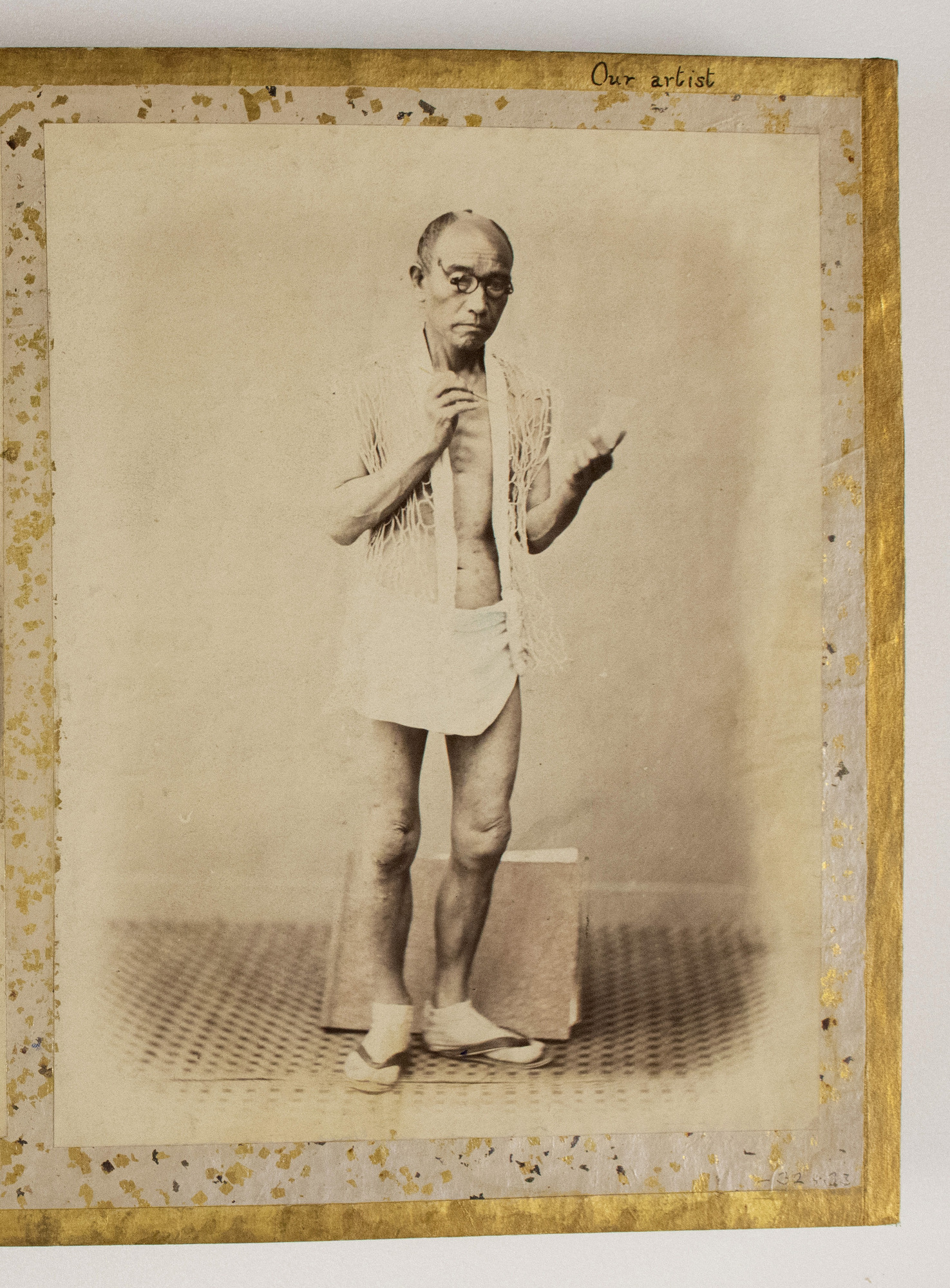 Sepia photograph colored with blue shades representing a standing half-naked Japanese man holding a brush and a small piece of paper, with an album behind his legs. He is photographed against a plain background. The photograph is placed in the center of an album page decorated with a gold-leaf frame, colorful dots, and an inscription.