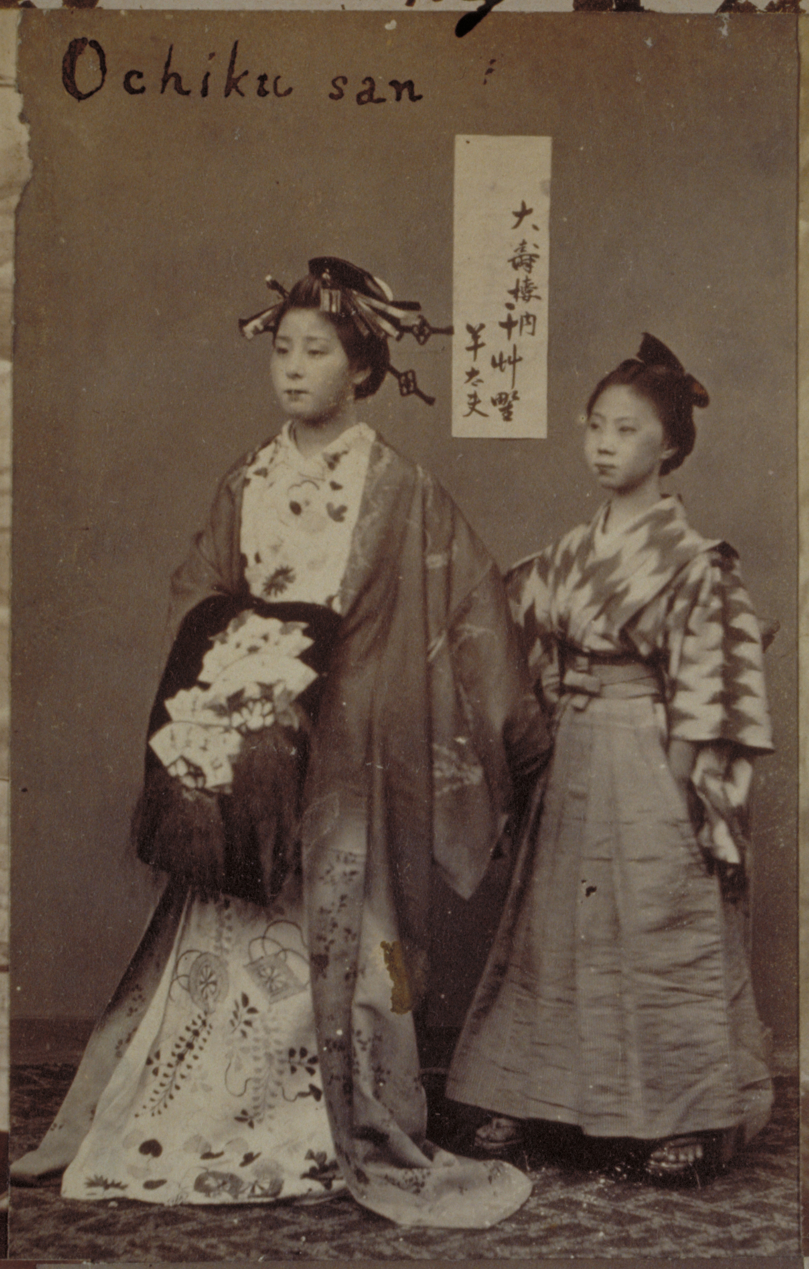 Black-and-white photograph with an inscription representing two Japanese women standing against a wall with a calligraphic inscription on it