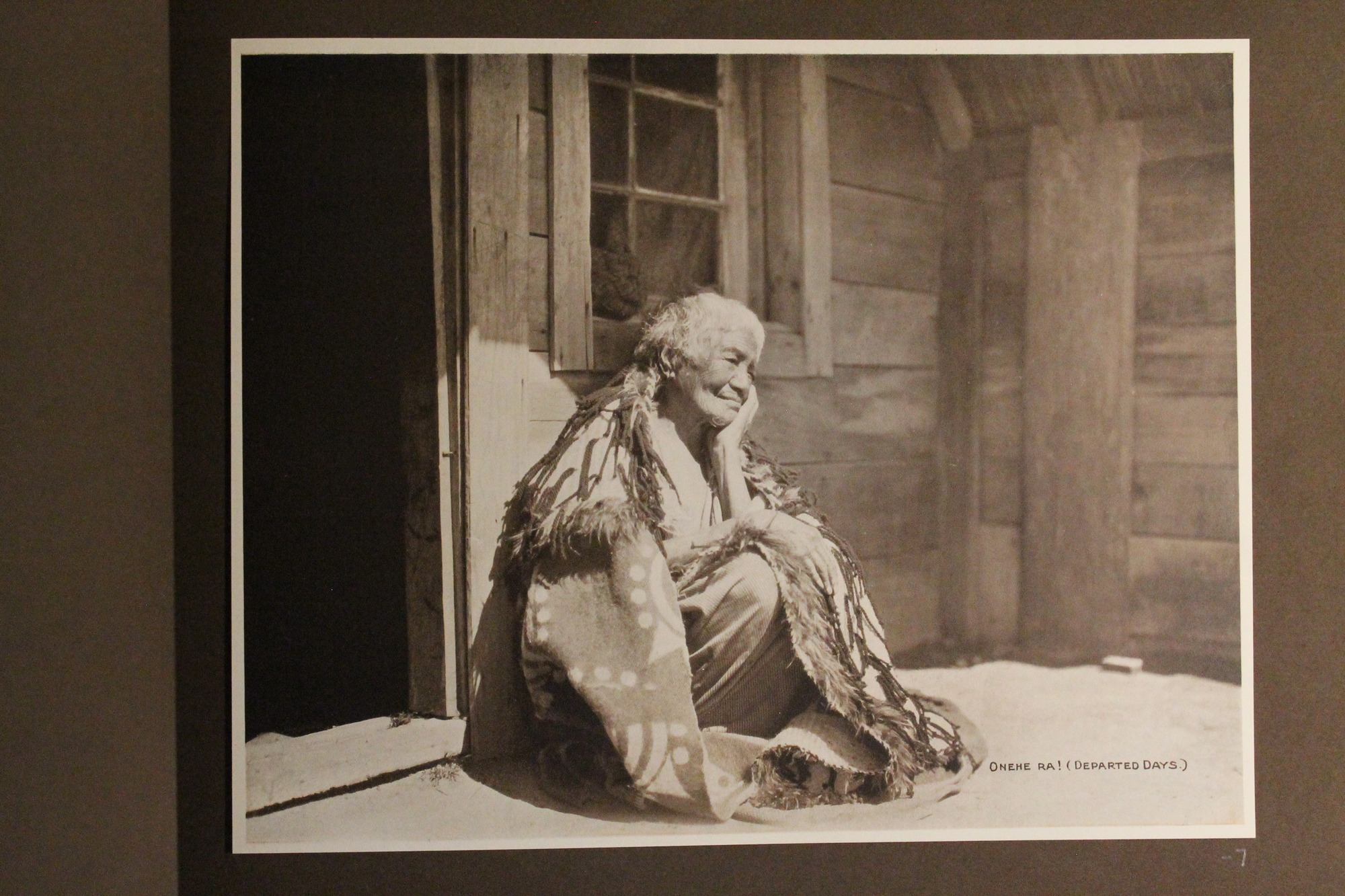 Old Maori woman sits outside wooden house. Black and white photograph.
