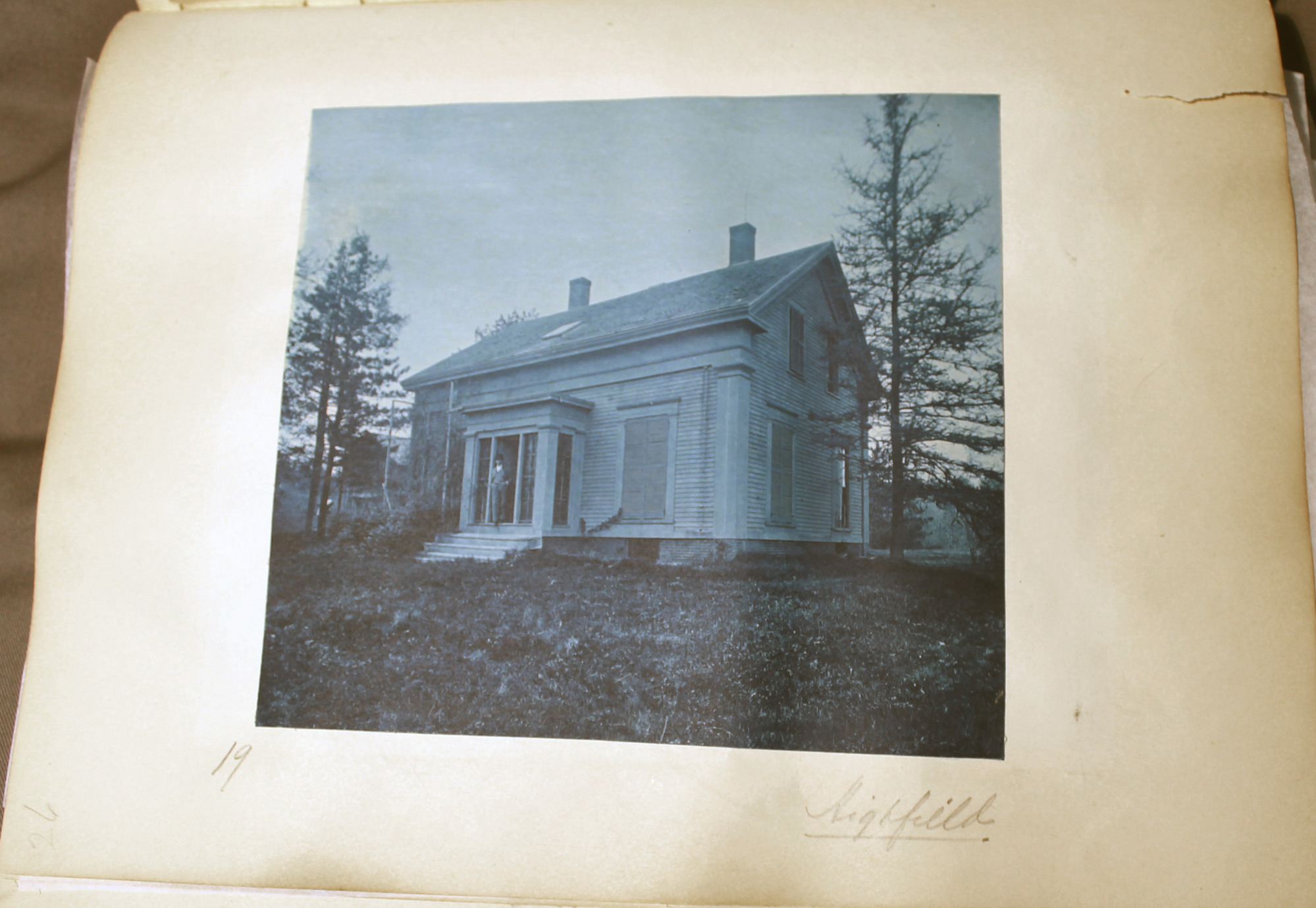 Blue toned photo of one-storied white clapboard building.  Man stands inside portico doorway.  Tall window shutters.  Evergreens on either side.