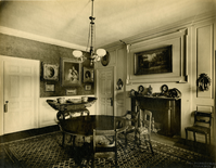 Black and white photograph of 19th century dining room featuring round dining table, fireplace and portraits of family members.