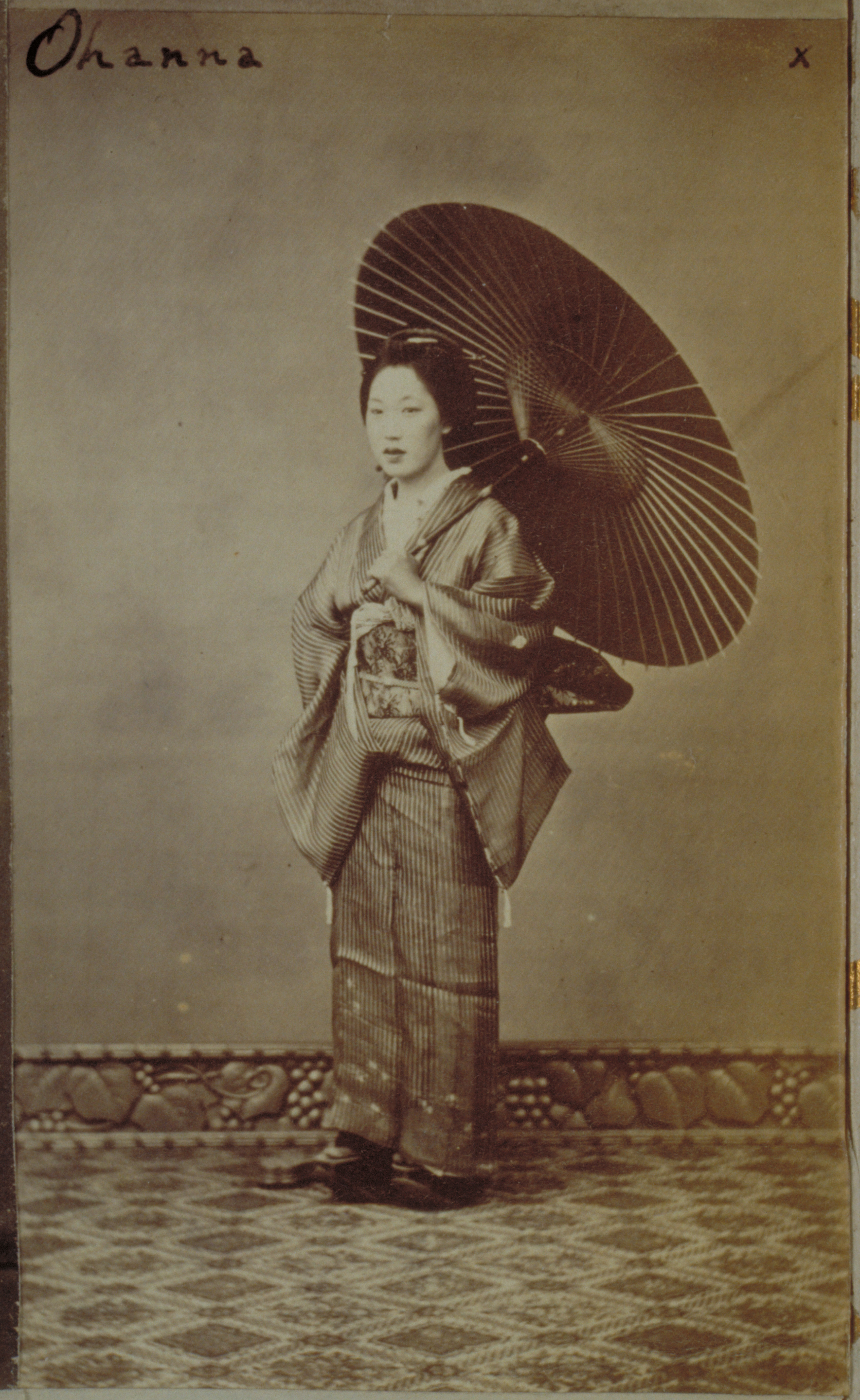 Black-and-white photograph with an inscription representing three-quarters of a Japanese woman standing, holding a parasol. Photographed by a plain wall with a carved molding along the floor.