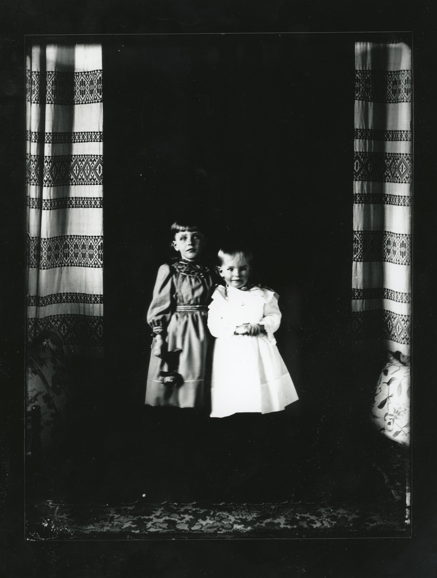 Two young white girls stand on a carpet in front of a background drape.