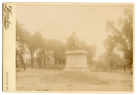 Photograph of a life-size or larger statue of Henry Wadsworth Longfellow. Statue is on a tall pedestal outdoors. Buildings in the background. Longfellow wears a suit and is draped in some kind of cloth, possibly a professor/graduation gown. He has chin-length hair and a full beard.