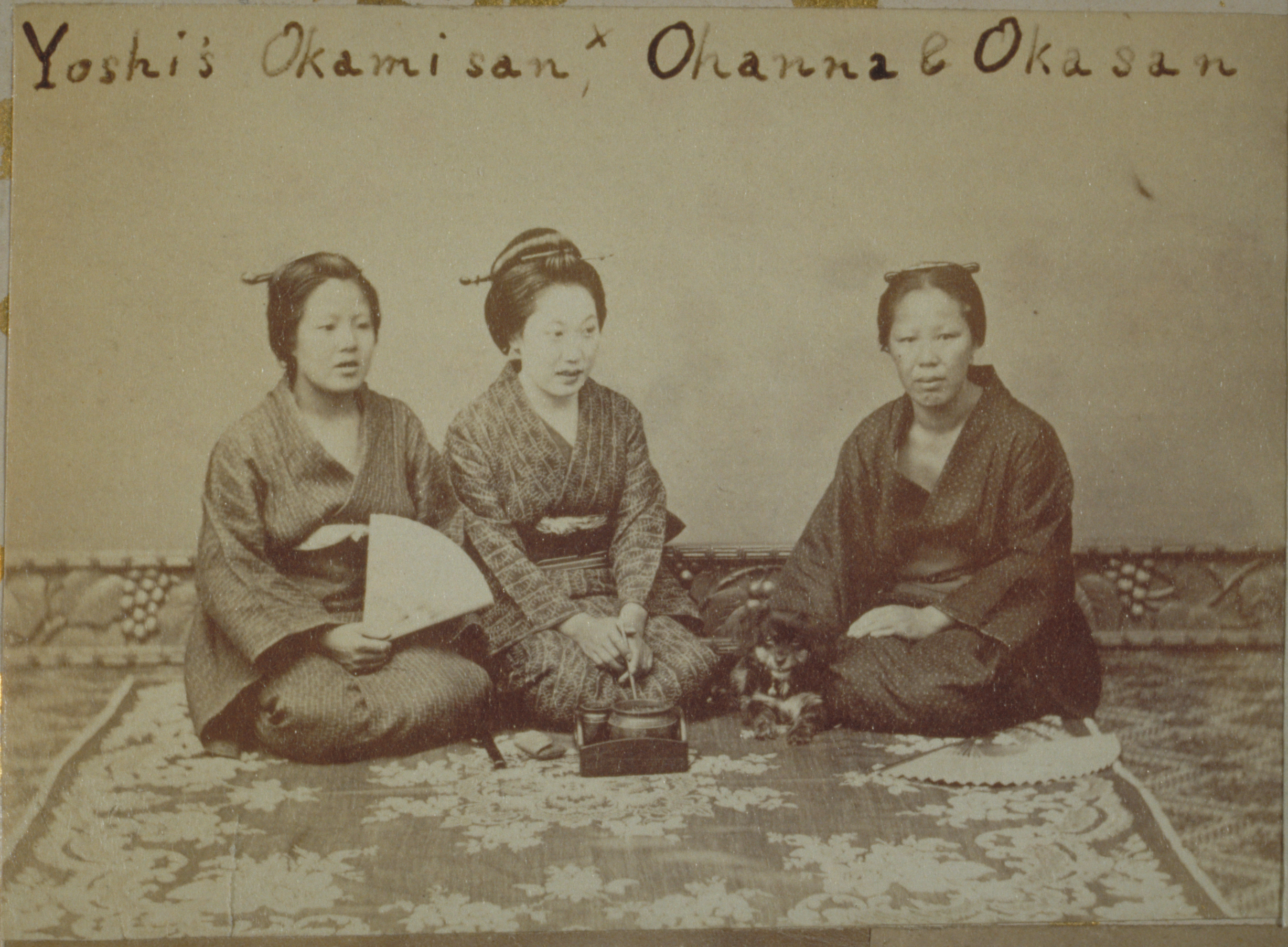 Black-and-white photograph with an inscription representing three Japanese women sitting on a rug, one holding a fan, one looking at the camera. They are photographed by a plain wall with a carved molding along the floor.