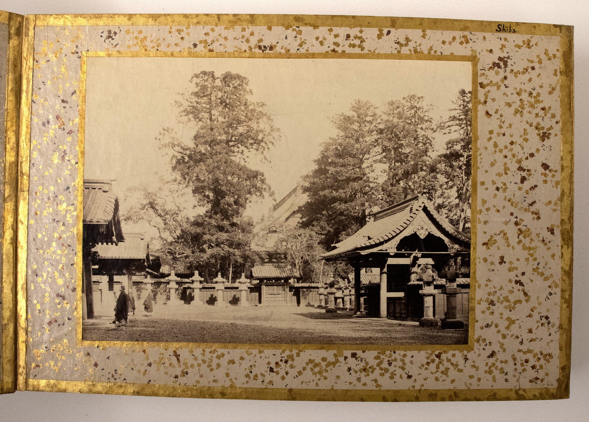 Sepia photograph representing a fence with stone lanterns along it and a complex of Japanese buildings behind the fence, trees in the background and a person in the front left. The photograph is placed in the center of an album page decorated with a gold-leaf frame, colorful dots, and an inscription.