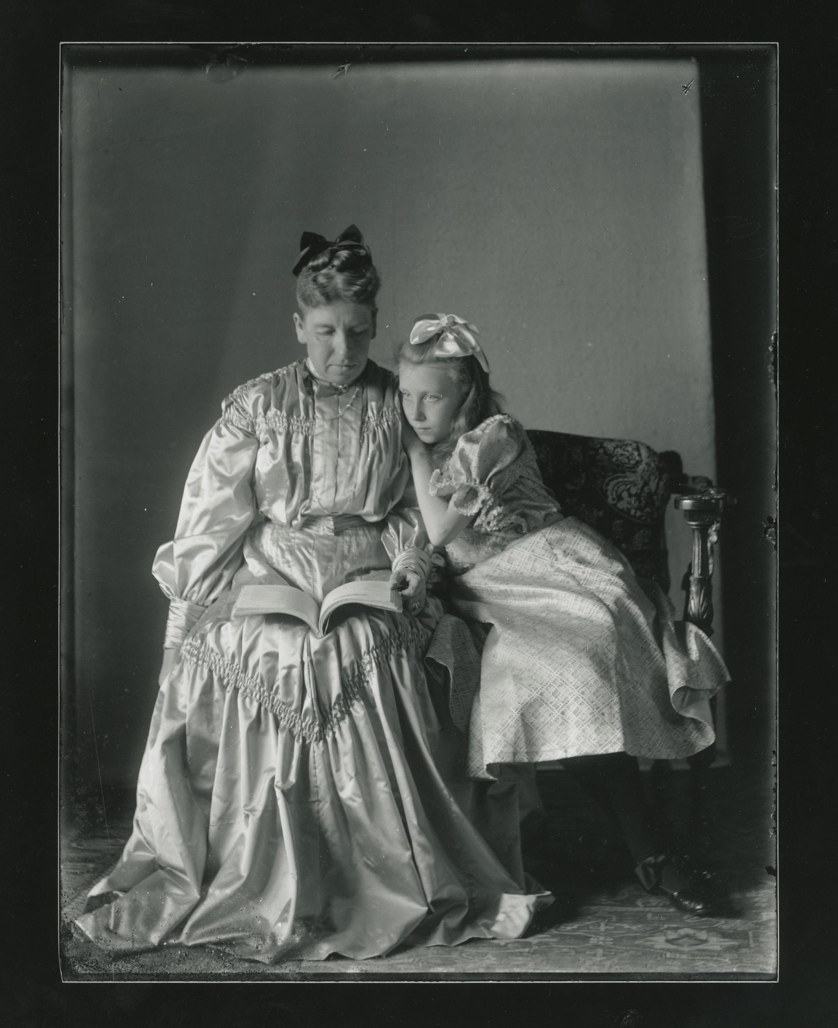 A white woman sits and reads on a loveseat next to a young girl.