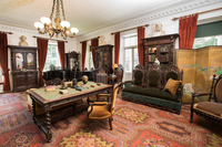Large room furnished with ornately carved wooden library table and bookcases, couch, and chairs.  Five large windows have red curtains.