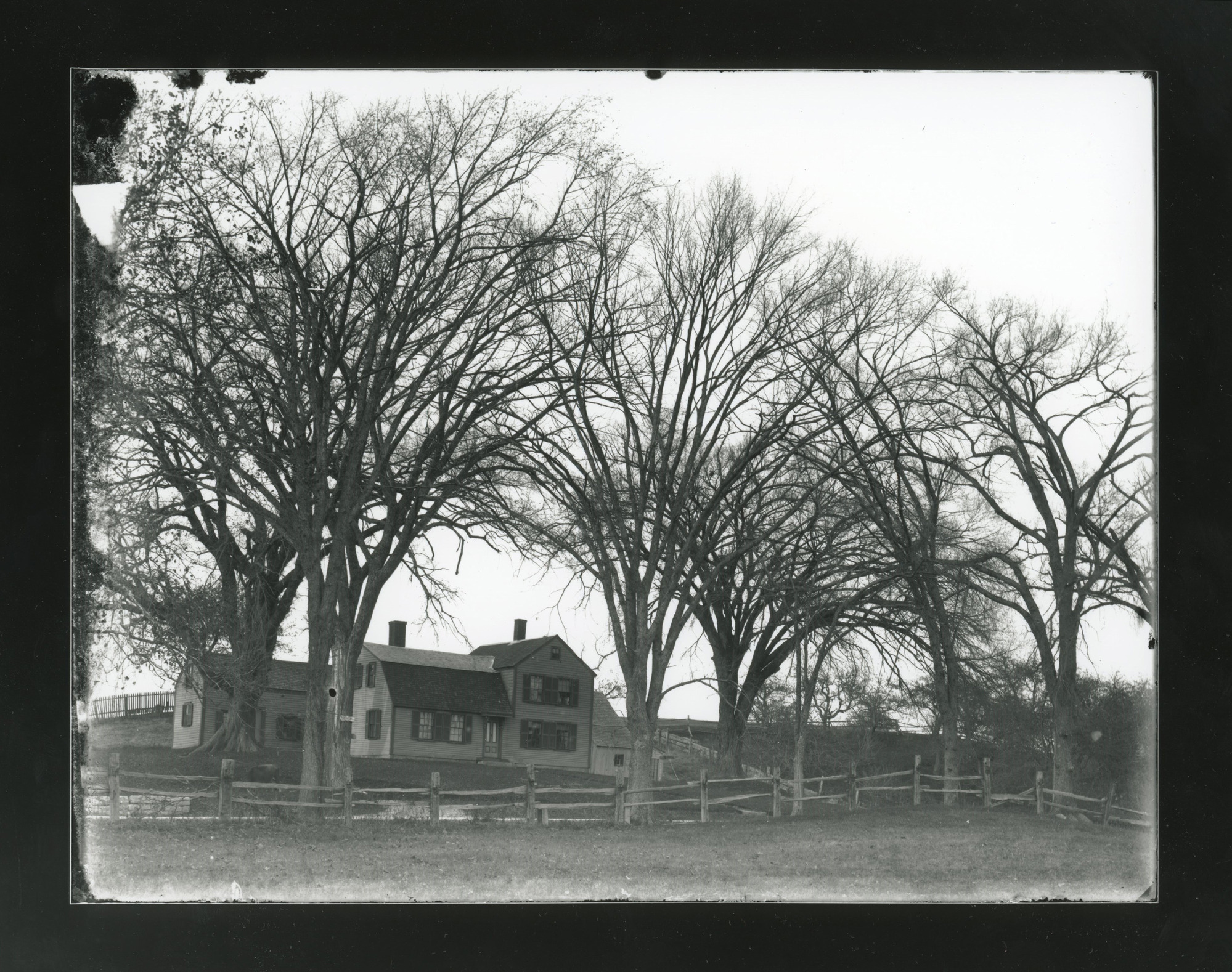 Photograph of a house and possibly a barn. Trees with no leaves are scattered around the house. Stone fence in the foreground.