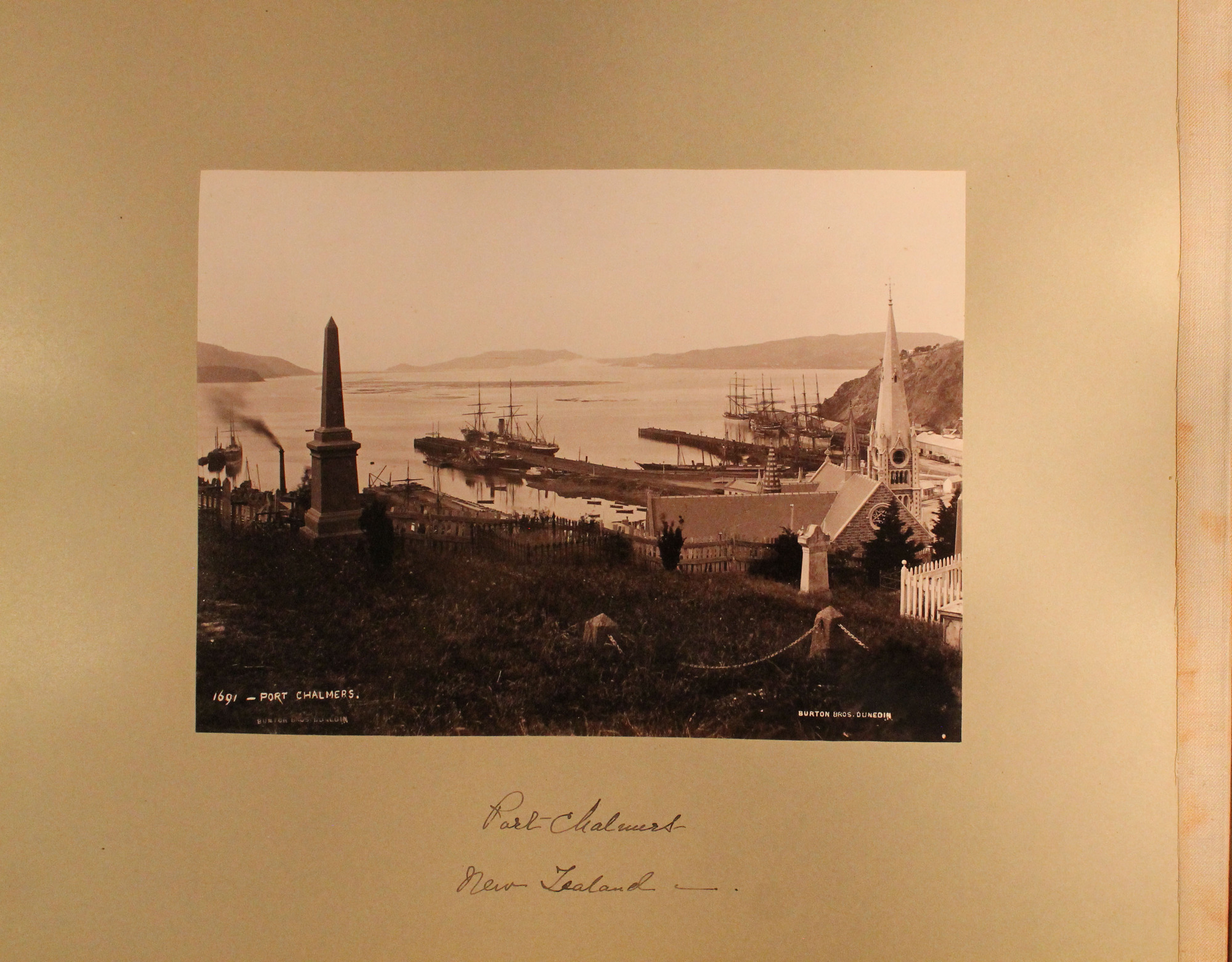 View of Port Chalmers, near Dunedin, seen from hilltop cemetery. Large ships alongside several long at docks. Presbyterian church in foreground, mountains in distance.