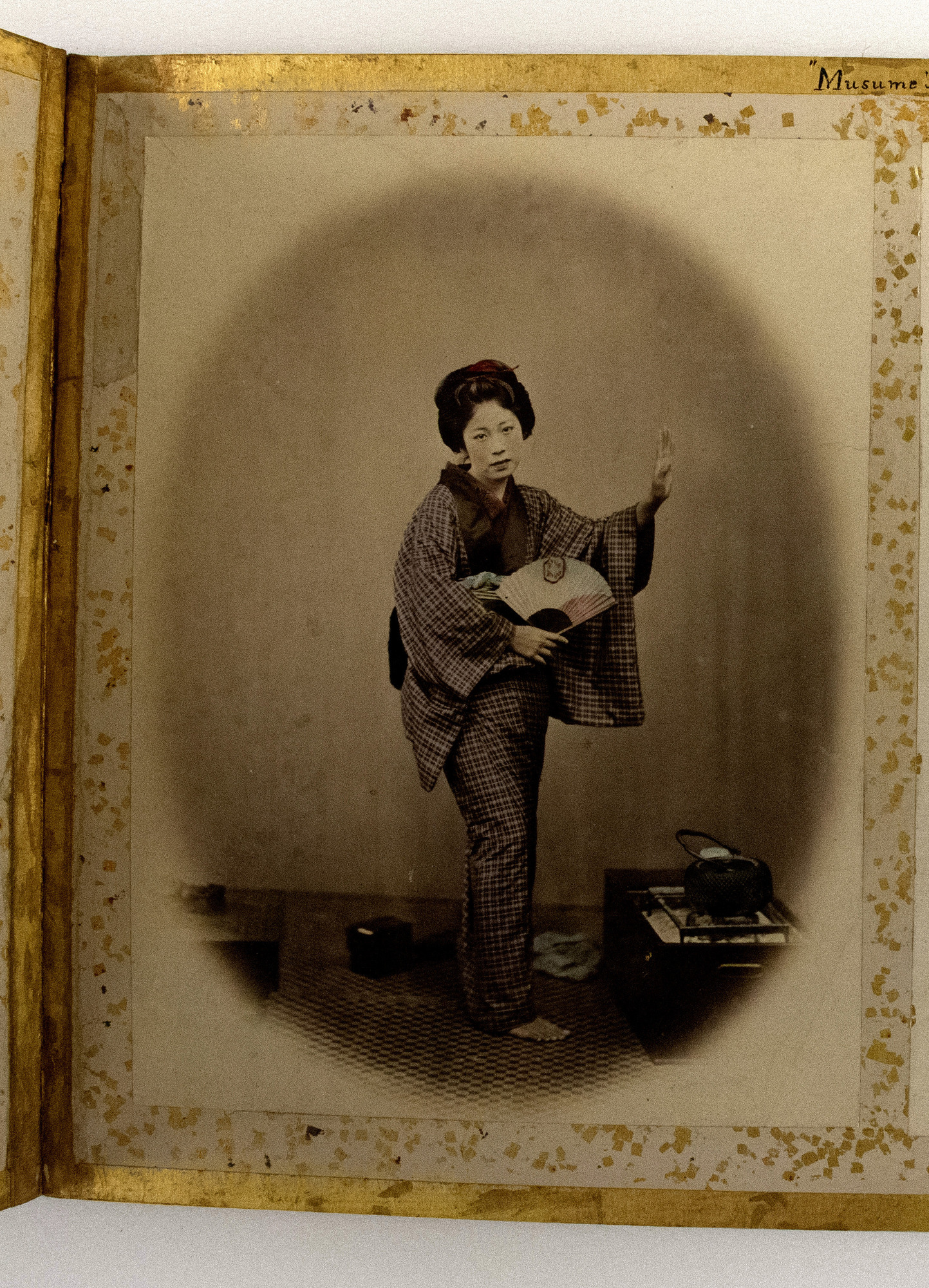 Sepia photograph colored with  blue, pink and red shades representing a Japanese woman standing on a mat and carrying a fan. They are photographed by a box, looking at the camera against a plain wall background. The photograph is placed in the center of an album page decorated with a gold-leaf frame, colorful dots, and an inscription.