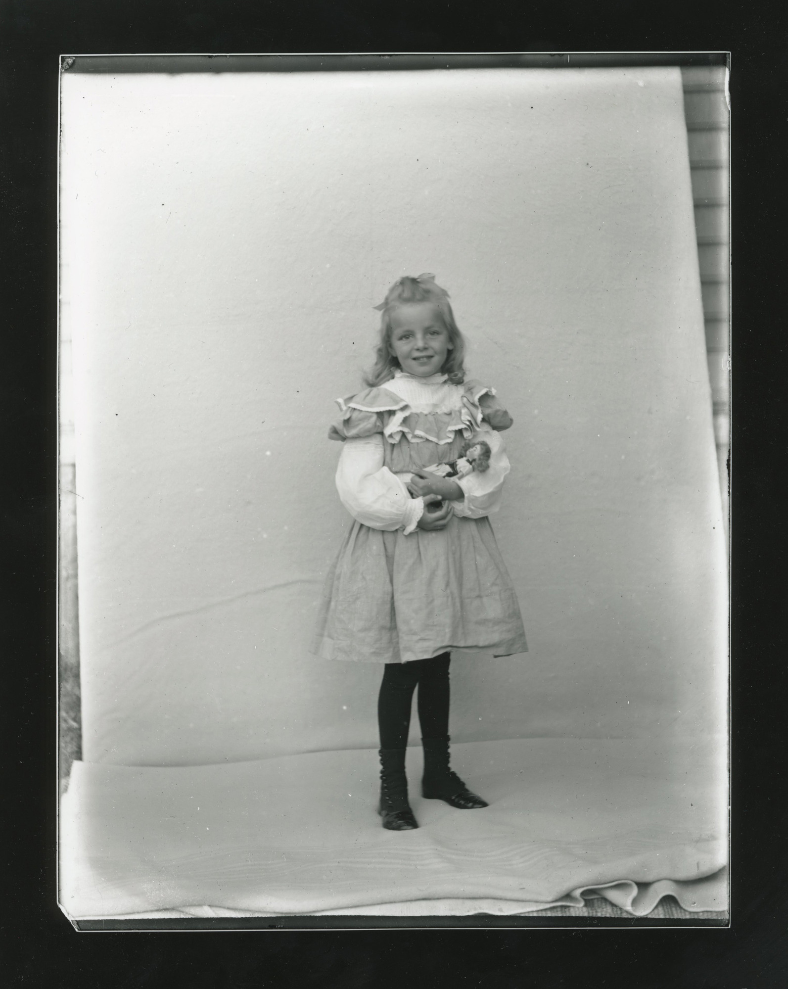 A young white girl stands in front of a backdrop. She holds a small white doll in her arms.