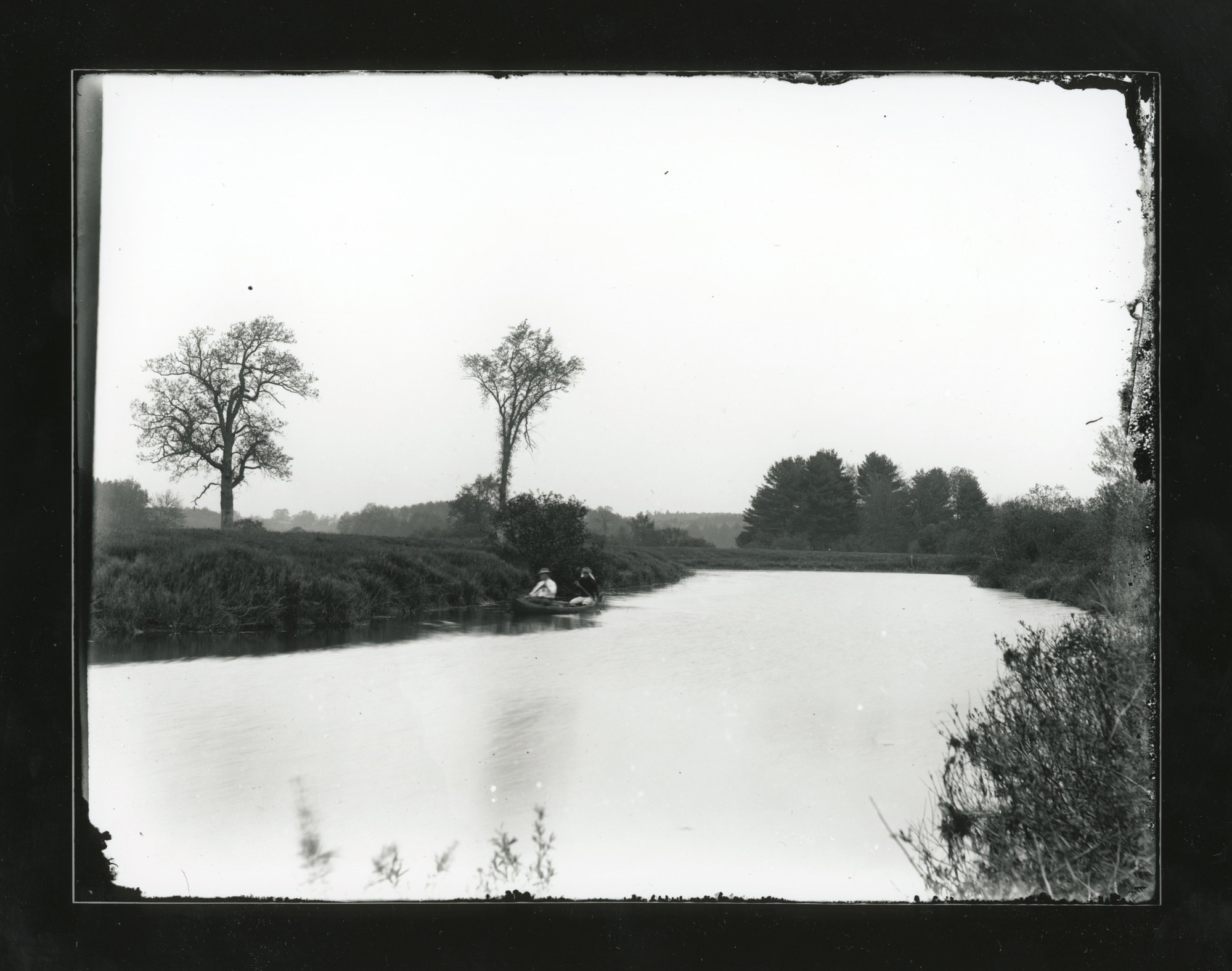 Two men in a canoe on a river. River is flanked on one side by a marsh and the other by trees.