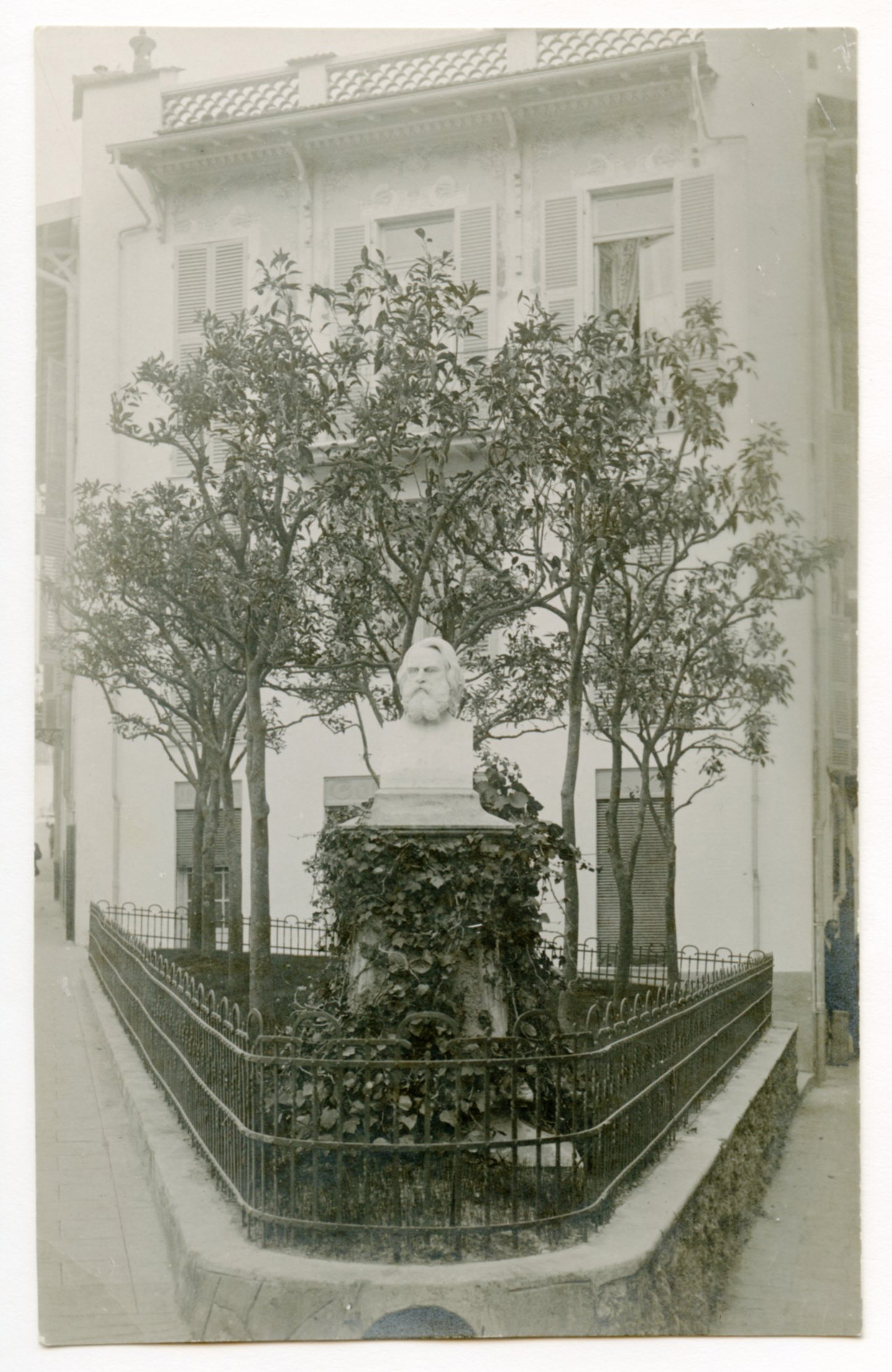 Henry Wadsworth Longfellow statue situated in triangular outdoor plaza. Plaza has grass and trees. Building in the background.