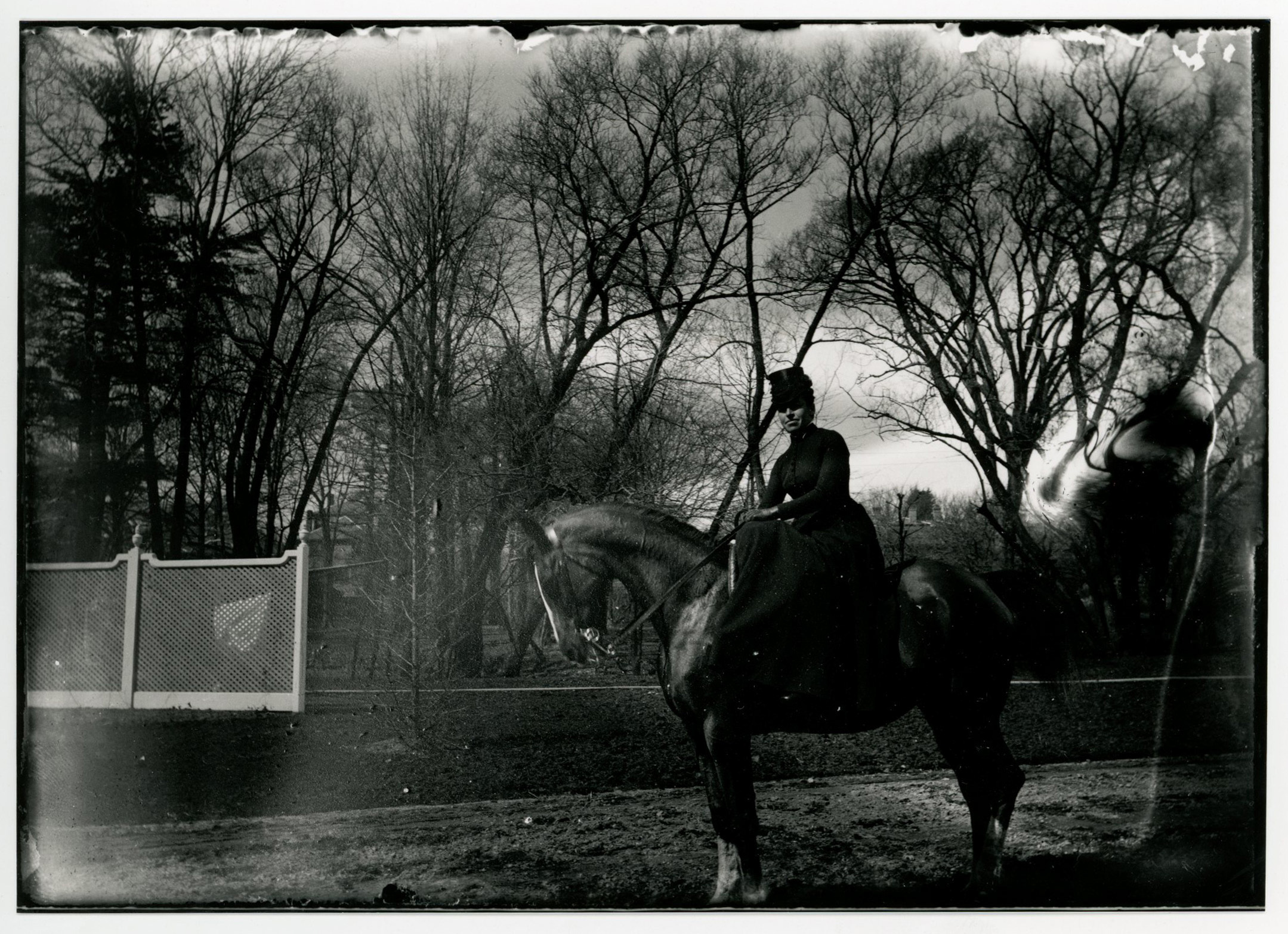 A white woman in riding hat and habit sits sidesaddle on a dark-colored horse. White fence in left midground. Trees in background.