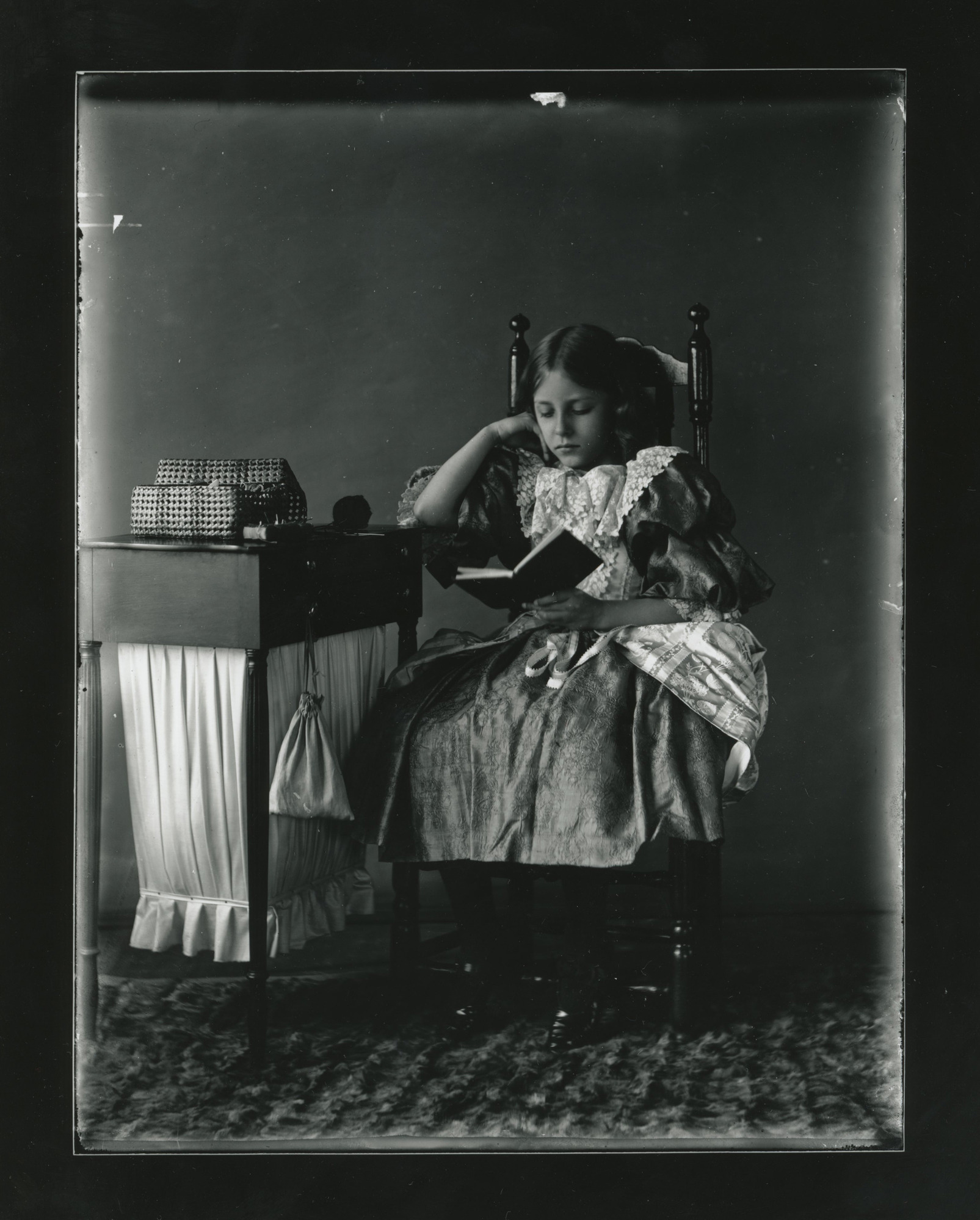 A young white girl sits in a chair and reads. She rests her right elbow on a side table with yarn and a sewing or knitting box.