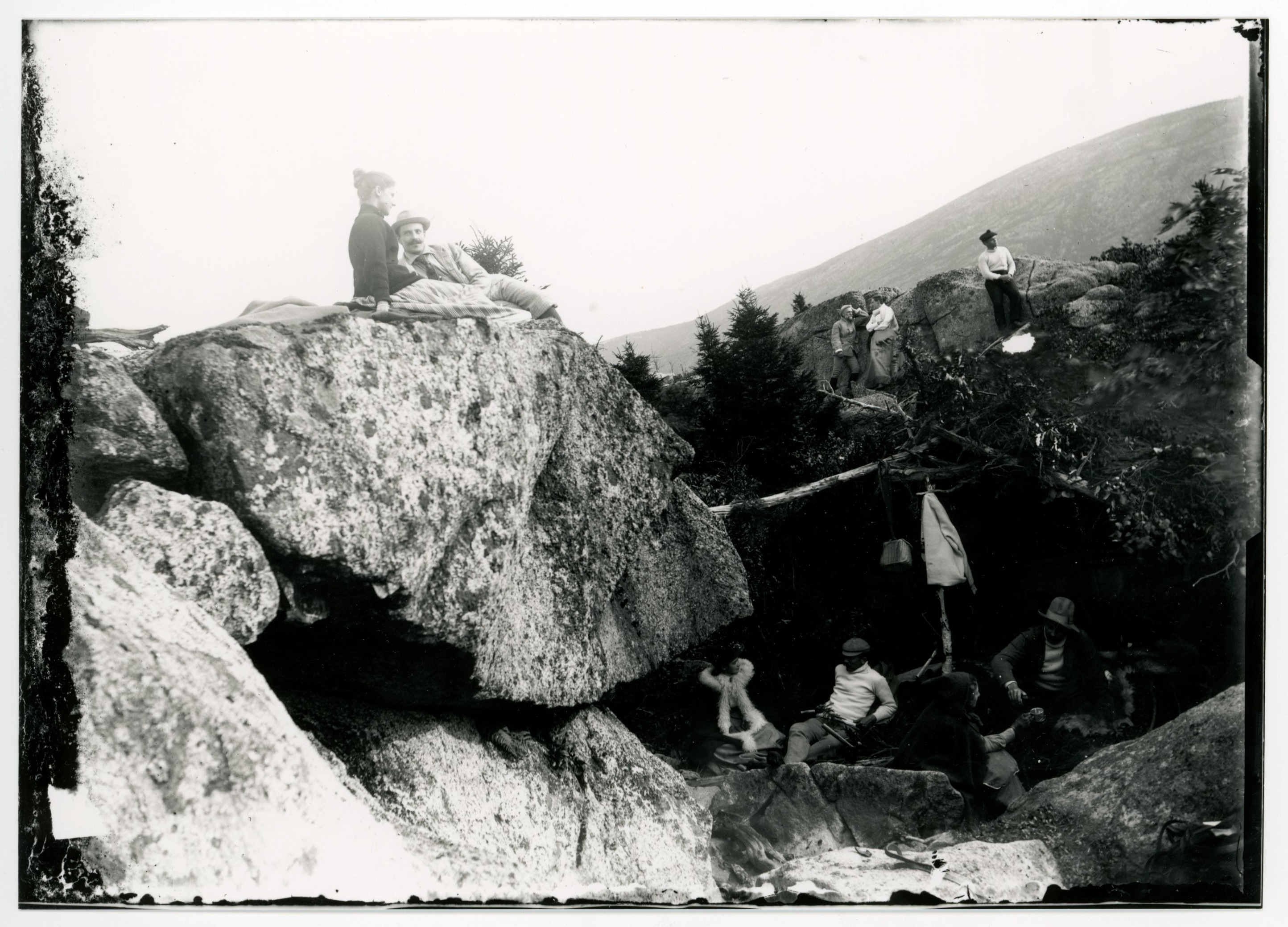 White men and women relax in between and on huge sections of rock. One man carries what seems to be a thermal carafe. A woman stands next to a small wicker basket. A man and a woman sit together on the highest rock. Evergreen trees are between the rocks, mountains in the background.