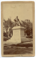 Photograph of a life-size or larger statue of Henry Wadsworth Longfellow. Statue is on a tall pedestal outdoors. Buildings in the background. Longfellow wears a suit and is draped in some kind of cloth, possibly a professor/graduation gown. He has chin-length hair and a full beard.