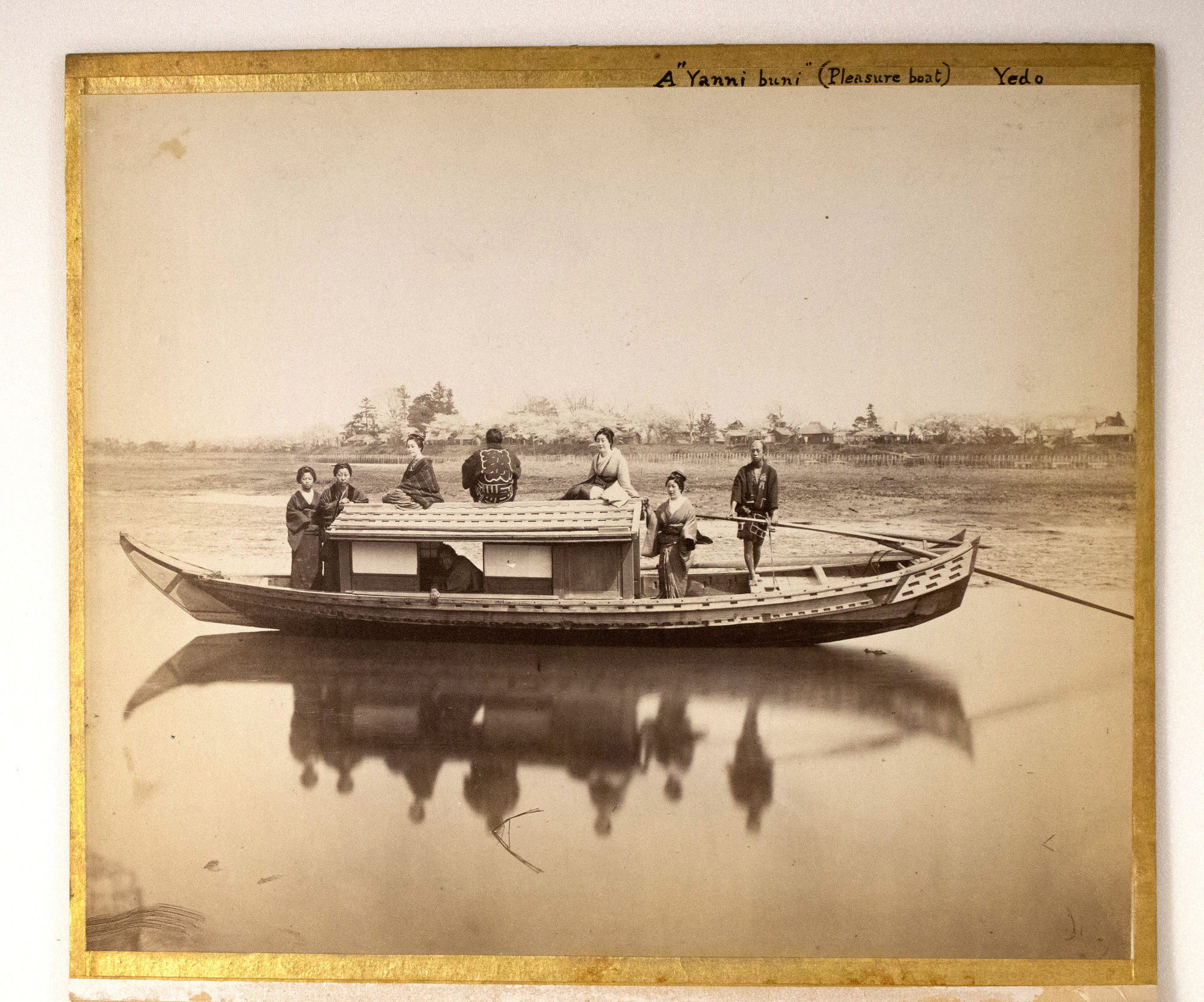 Black-and-white photograph representing eight Japanese women and men, sitting and standing on a boat in the river with a landscape behind them.