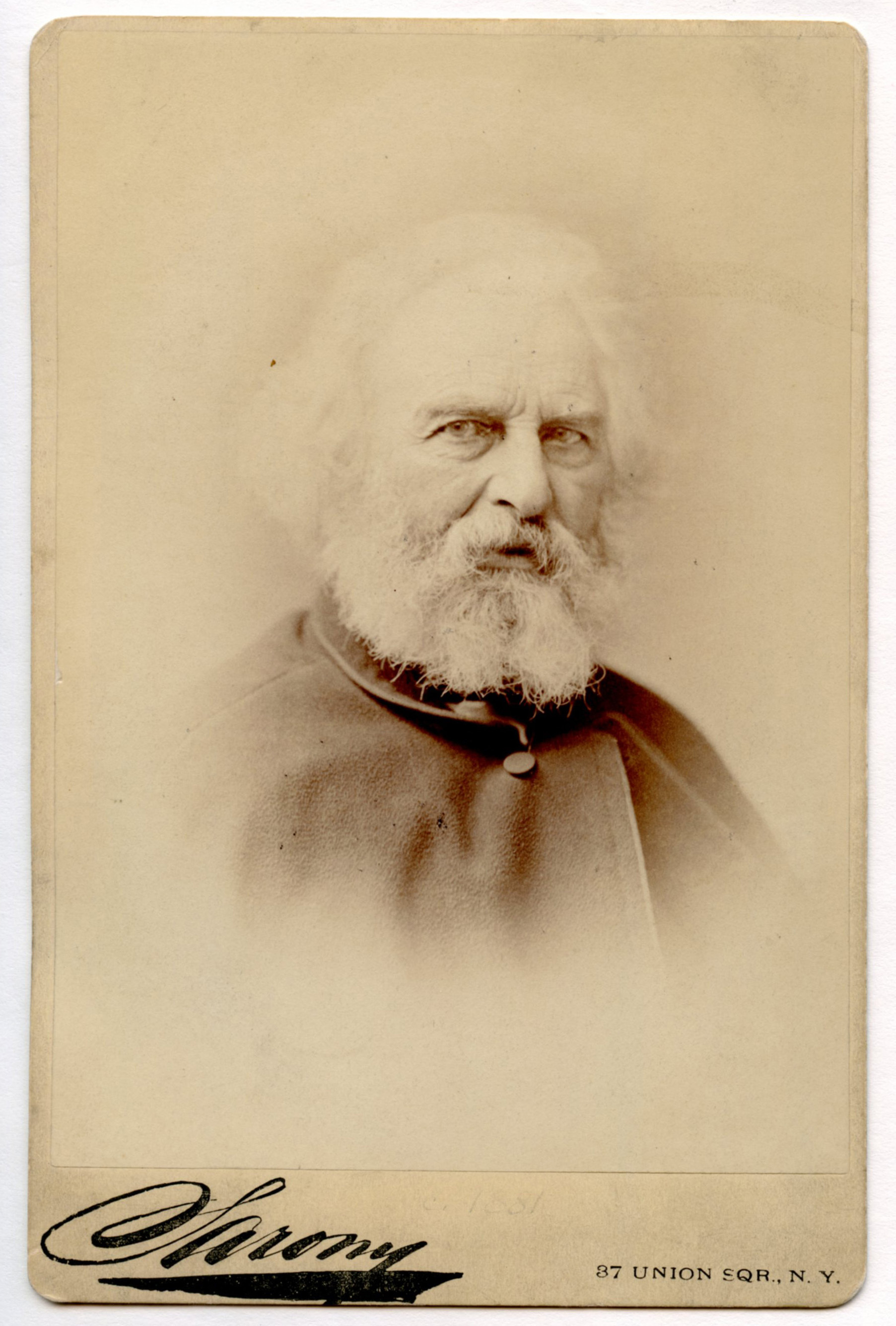Henry Wadsworth Longfellow dressed in a buttoned cloak. He looks almost directly at the viewer. Head and shoulders visible against blank background. Photograph has a vignette effect. Longfellow has chin-length white hair and a full white beard.