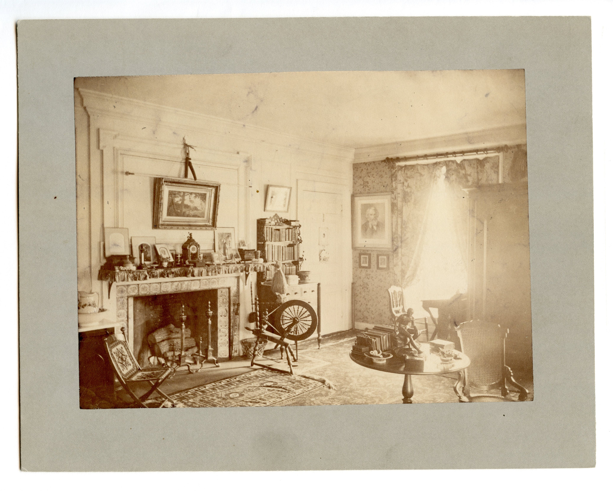 Black and white photograph of Victorian bedroom with fireplace, spinning wheel and bookshelf featured.