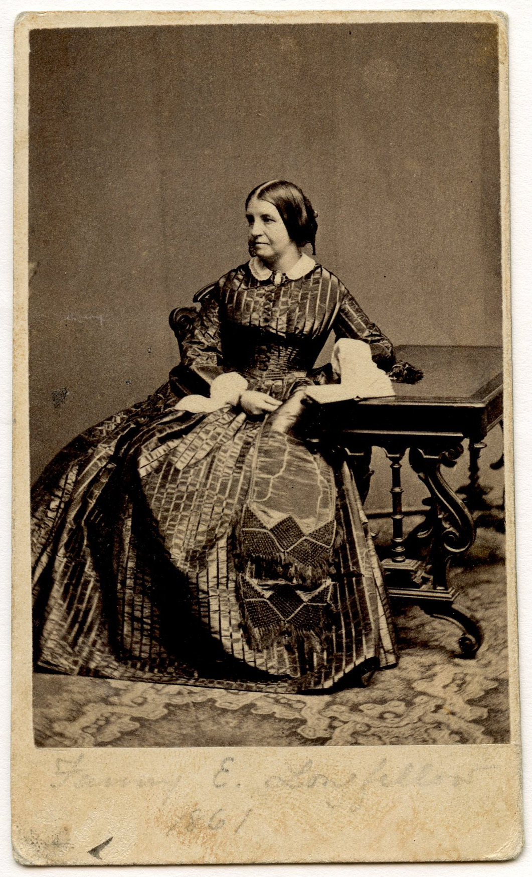 A portrait of a white woman sitting with her left hand on a book. The book is placed on the Renaissance Revival style table to her side. A patterned carpet is visible. She wears a striped dark-colored dress with white collar and undersleeves.