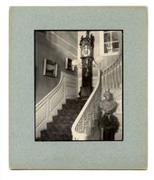 Black and white photograph of grandfather clock at the top of stairs.