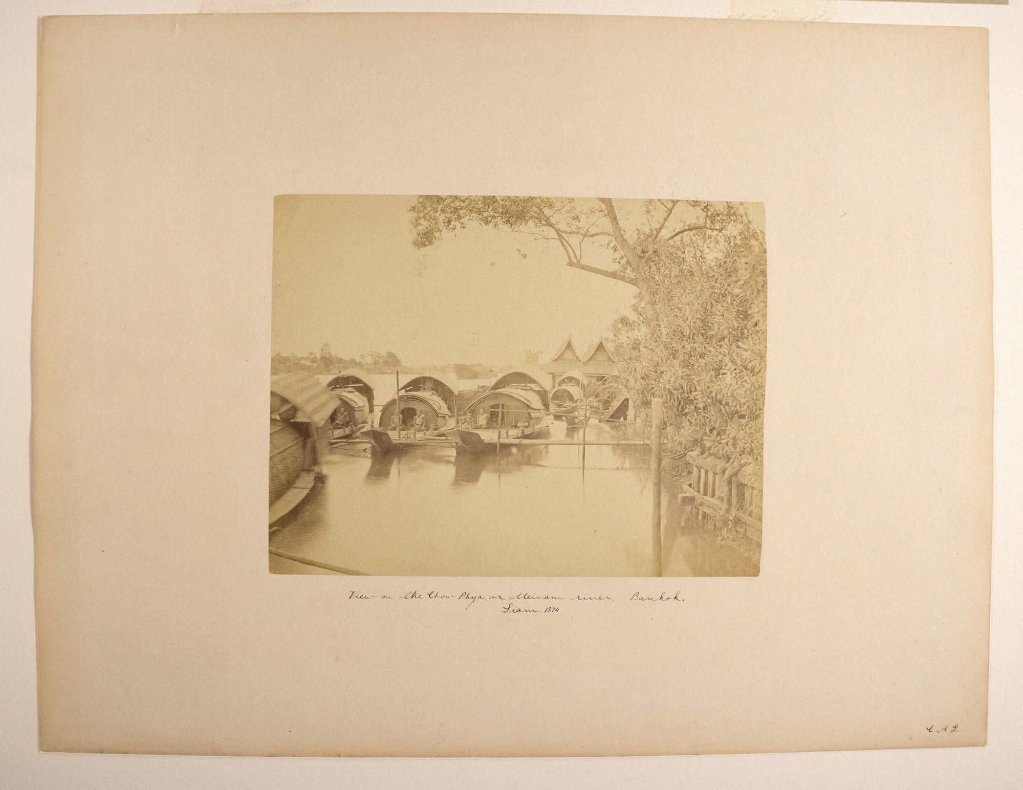Sepia photograph with an inscription representing several houseboats on water, people sitting by entrances, and plants on the right.