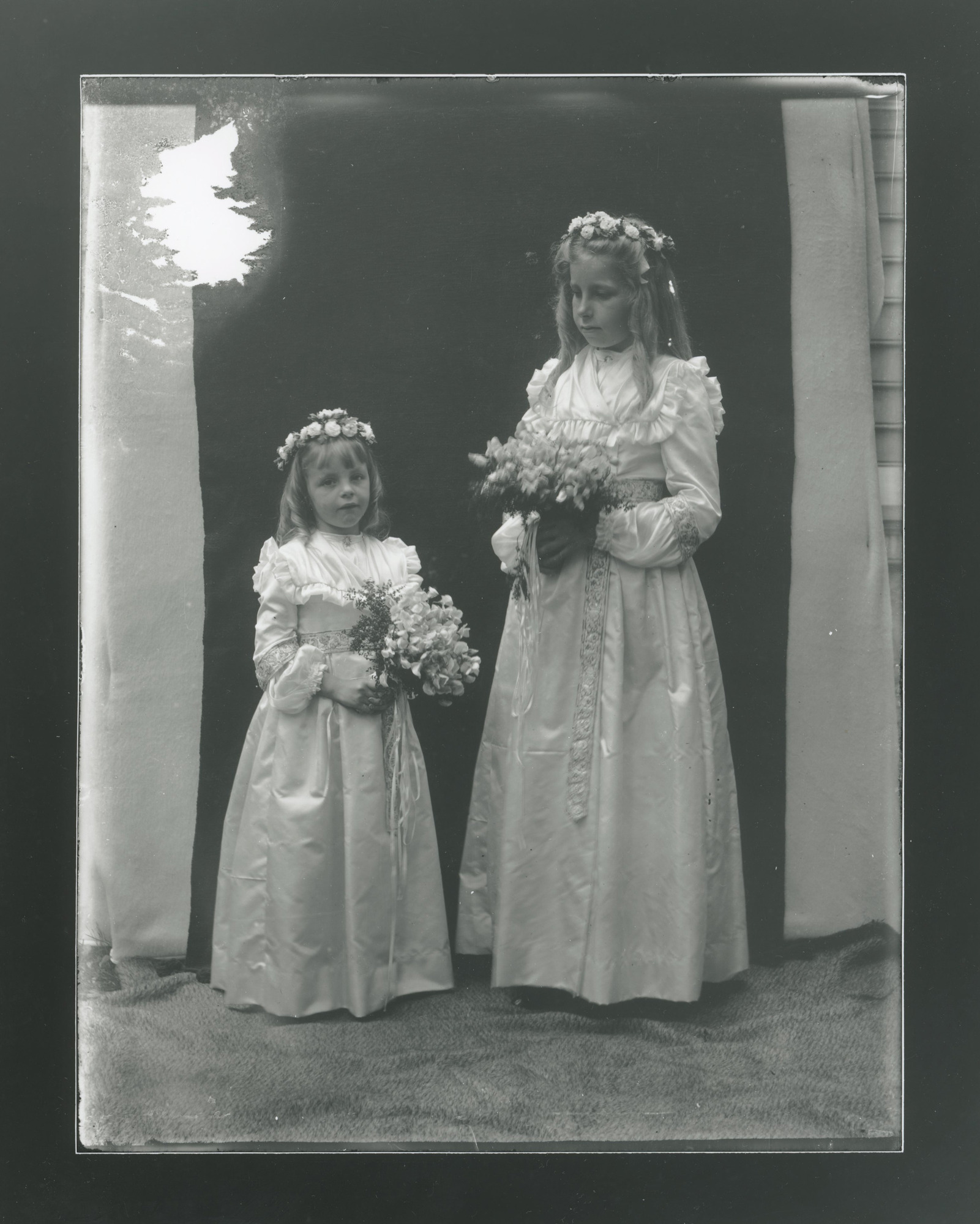 Two white children in ruffled white dresses hold hands. In free hand they each hold a bouquet of flowers. Both wear flower crowns.