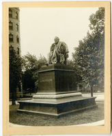 Photograph of an approximately life-size statue of Henry Wadsworth Longfellow on a tall pedestal outdoors. Longfellow wears a suit and a professor/graduation gown. He has chin-length hair and a full beard. He leans his chin on his right hand and holds a book casually in his left, his index finger keeping his page for him. In the left background is a tall building behind trees and in the right background is a short building behind trees.