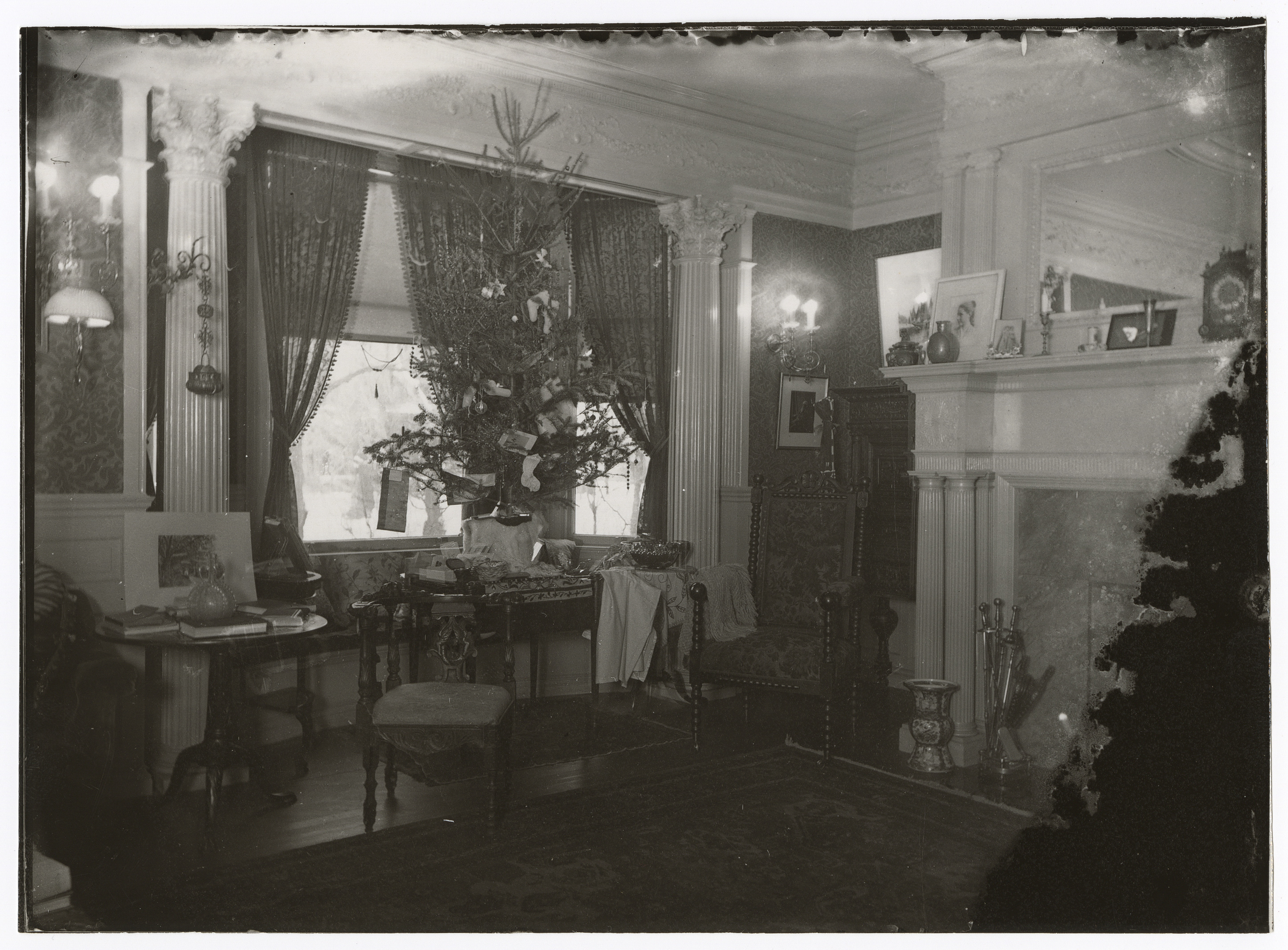 Christmas tree on a table in front of two windows. Cluttered side-table at left. Fireplace at right.