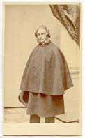 Henry Wadsworth Longfellow wearing a coat with short cape is shown standing at full length. The coat hides his arms and hands. A top hat to his right side is visible. The studio background includes a wooden square column, part of a brocade drape, and floor covering with alternating squares of light and dark. Longfellow has ear-length hair and sideburns.