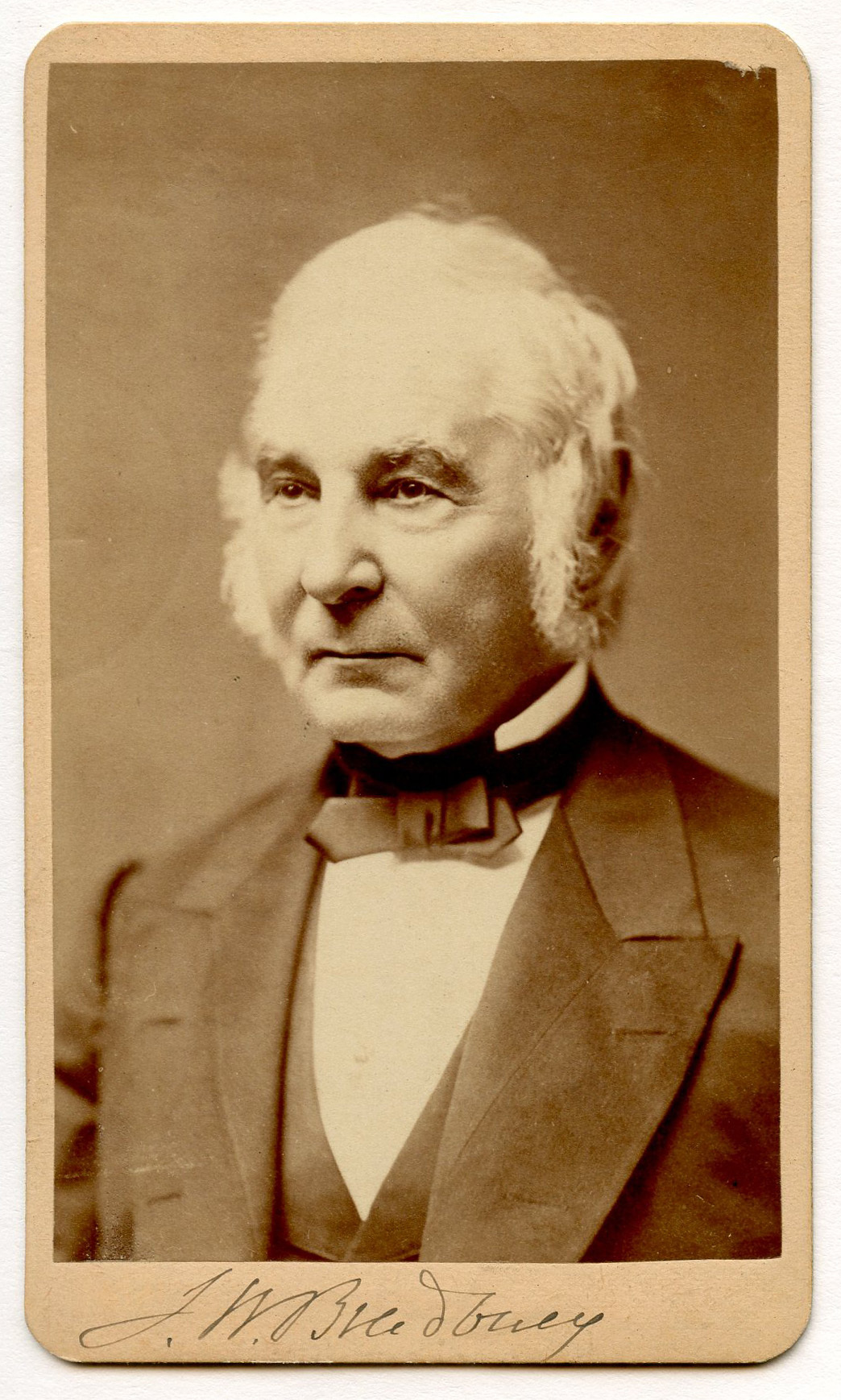 Portrait of a white man in three-quarter view to his right. He has white hair and sideburns. Two buttonholes are evident on each of his jacket's lapels. He is wearing dark formal attire and a bow-tie over a white collar.
