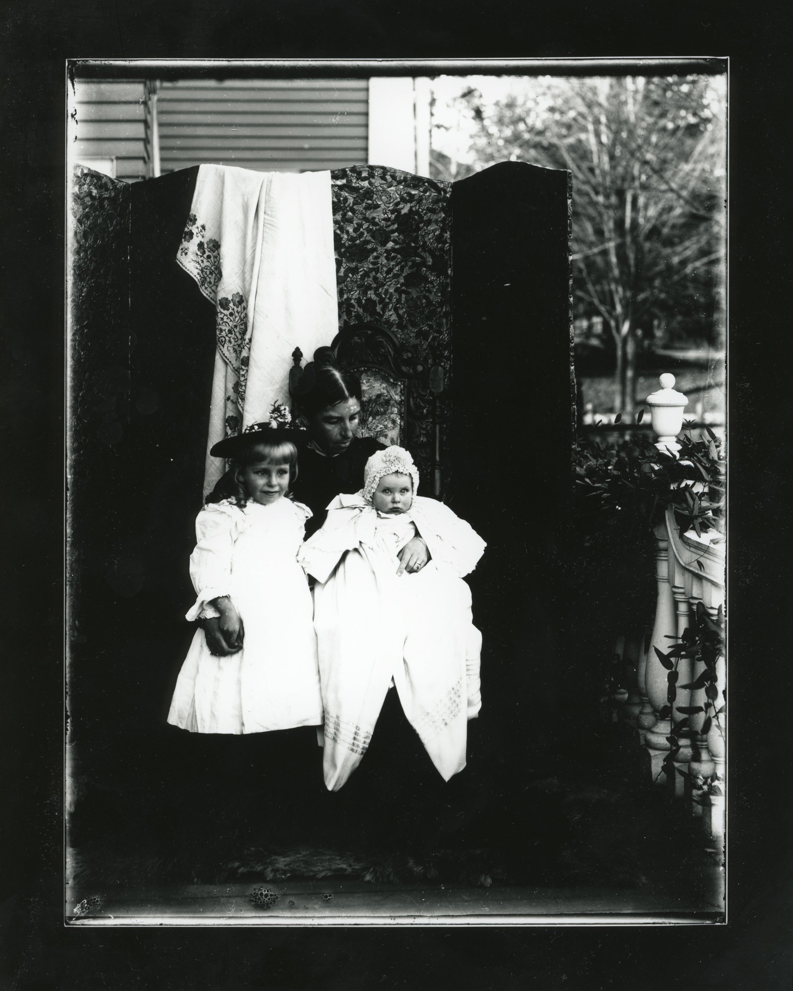 A white woman sits on a chair outdoors holding a baby on her left knee and putting her right arm around a young girl standing next to her. They pose in front of a room divider with a floral design.