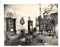 Black and white photograph of 19th century study with round table, fire place and several portraits in view.