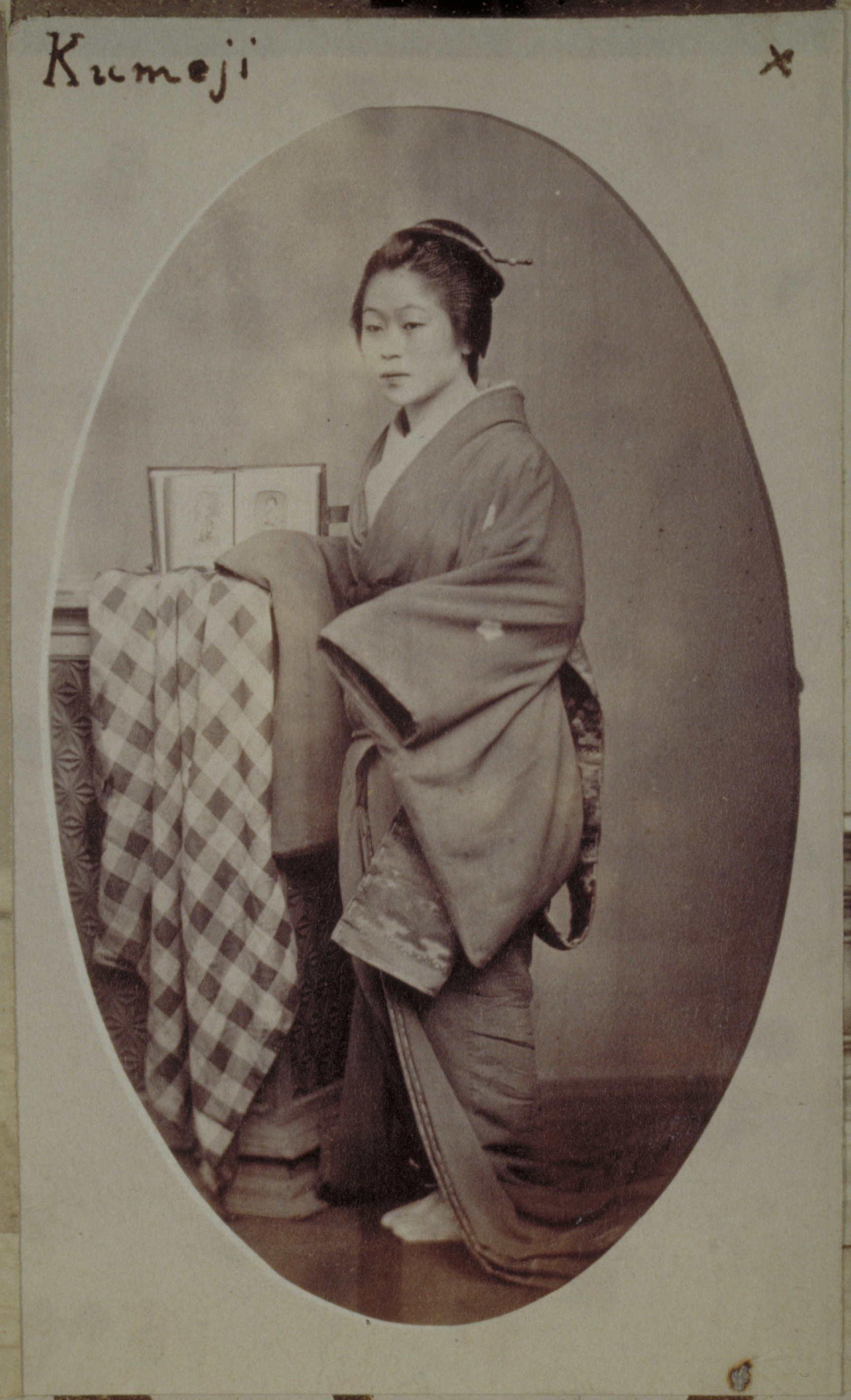 Oval shaped black-and-white photograph with an inscription representing a Japanese woman standing against a short column with draped cloth and an open book on it. She is photographed against a plain wall background.