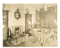 Black and white photograph of 19th century study. Fireplace, three bookshelves and circular desk featured.