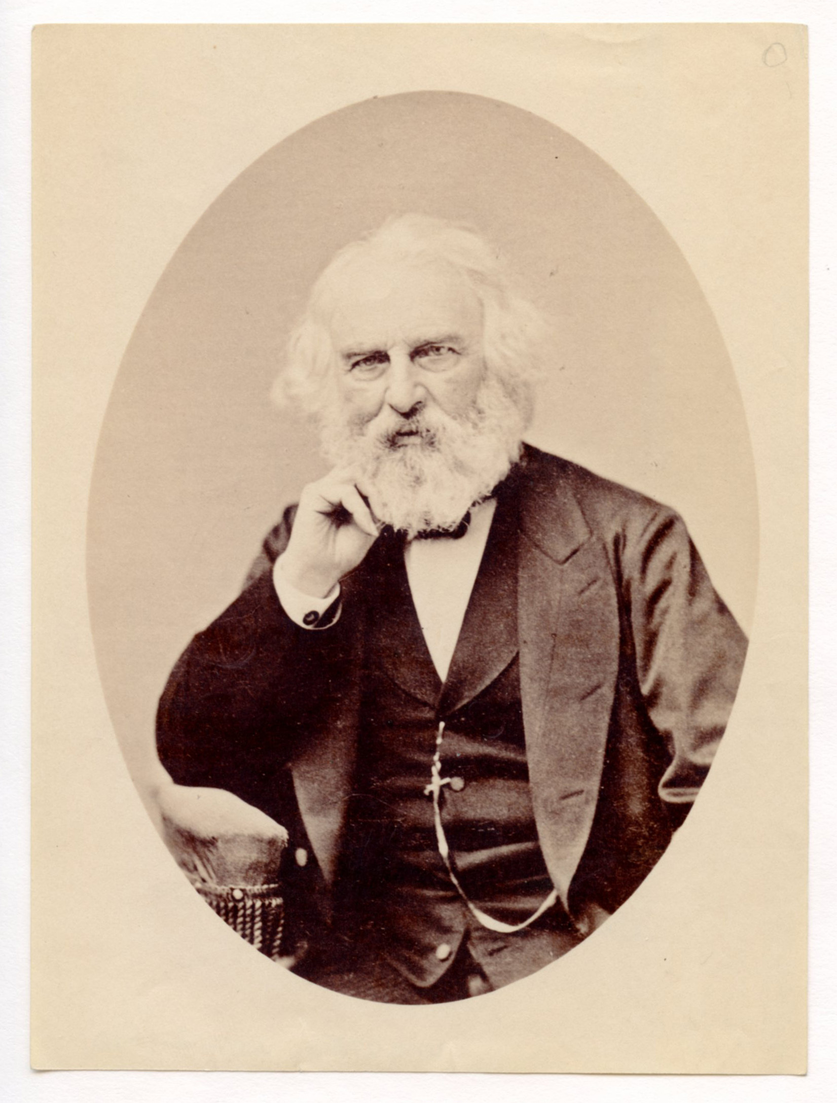 Henry Wadsworth Longfellow seated with his right elbow resting on the arm of his chair and his right hand supporting his chin. The upholstered arm of the chair with fringe is visible to the lower left. Longfellow wears a dark suit and has ear-length white hair and a full white beard.